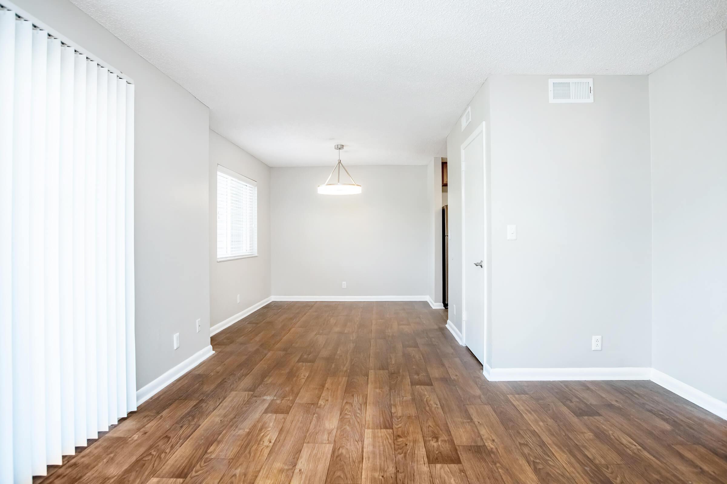 A spacious, light-colored living area with wooden flooring. The room features a window with vertical blinds, a hanging light fixture, and a door leading to another space. The walls are painted in a neutral tone, creating a clean and inviting atmosphere.