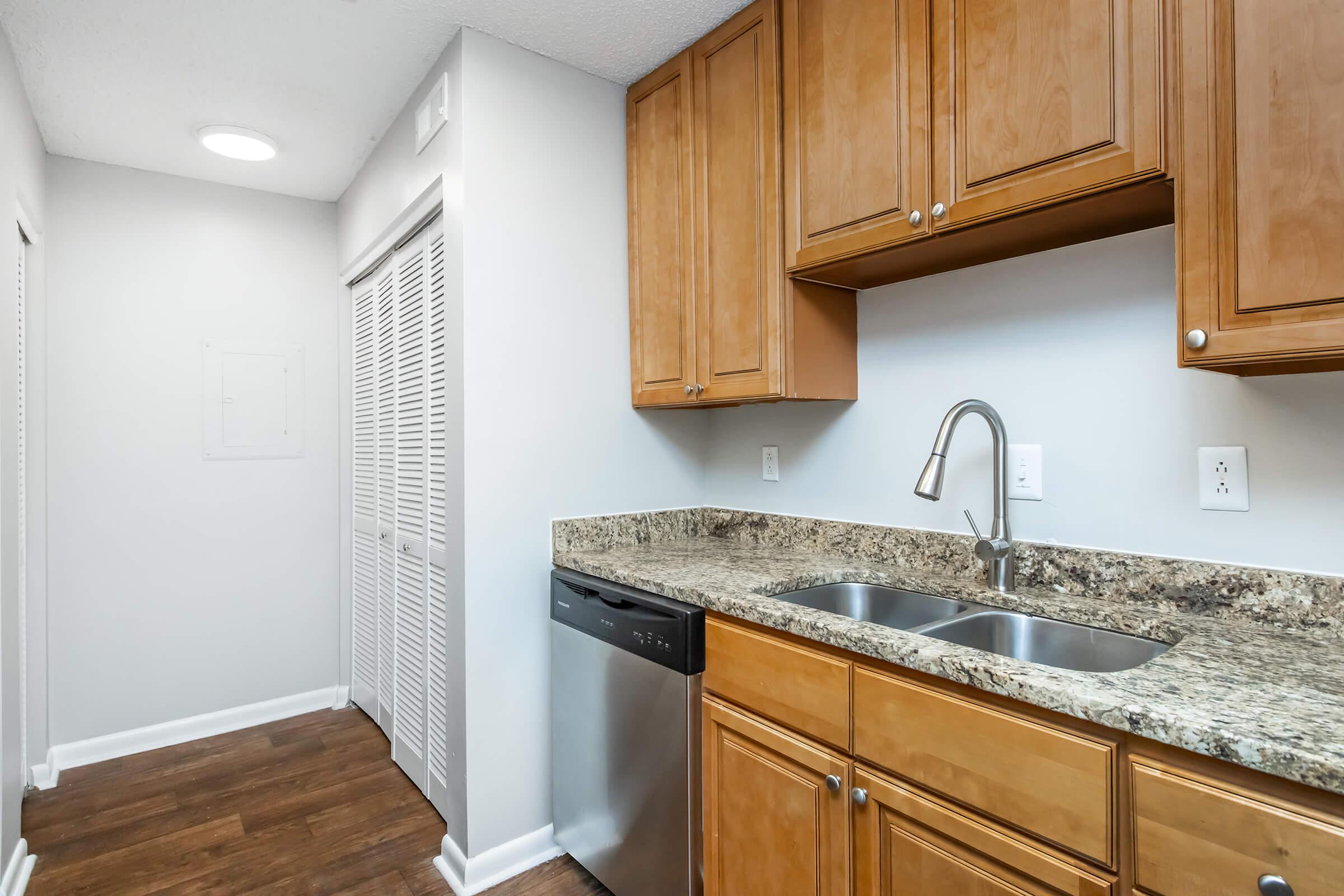 A modern kitchen featuring wooden cabinets, a granite countertop, double sink, and a stainless steel dishwasher. The space has light gray walls and a laminate wood floor. Adjacent to the kitchen is a hallway with sliding closet doors. Bright, well-lit environment.