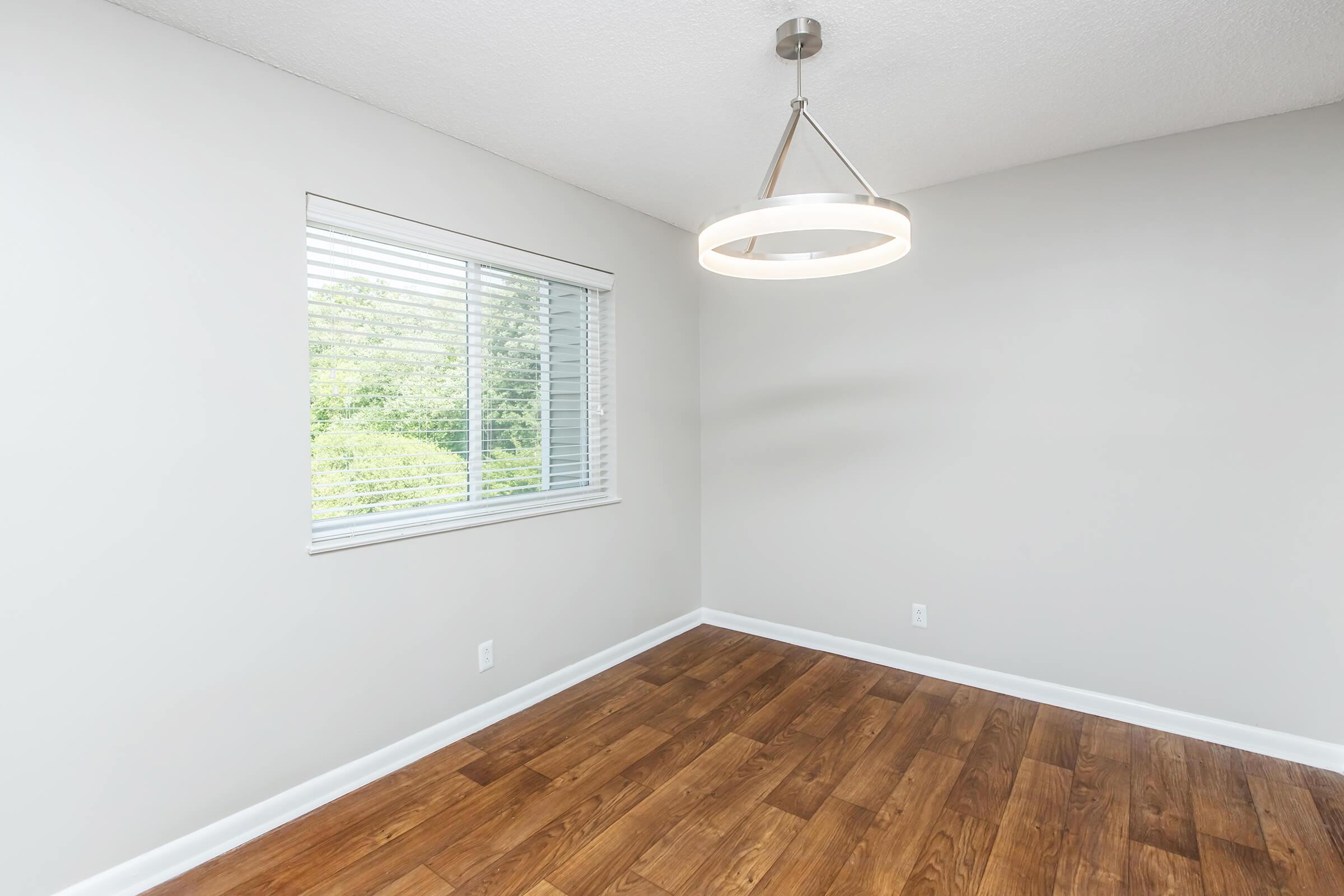 A bright, empty room featuring a modern pendant light fixture. A large window with blinds allows natural light to flood in, illuminating the light-colored walls and the warm wooden flooring, creating a spacious and inviting ambiance.