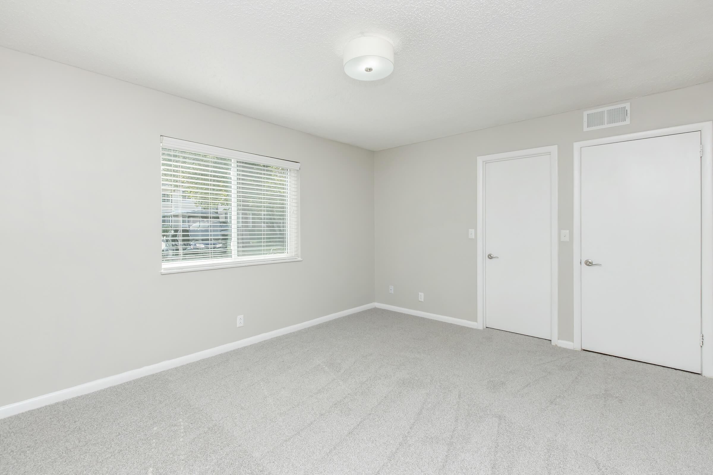 A bright, empty room with light-colored walls and carpet. It features a window with blinds, allowing natural light to enter. There are two doors on opposite walls, one of which appears to lead to a closet. The ceiling has a simple light fixture.