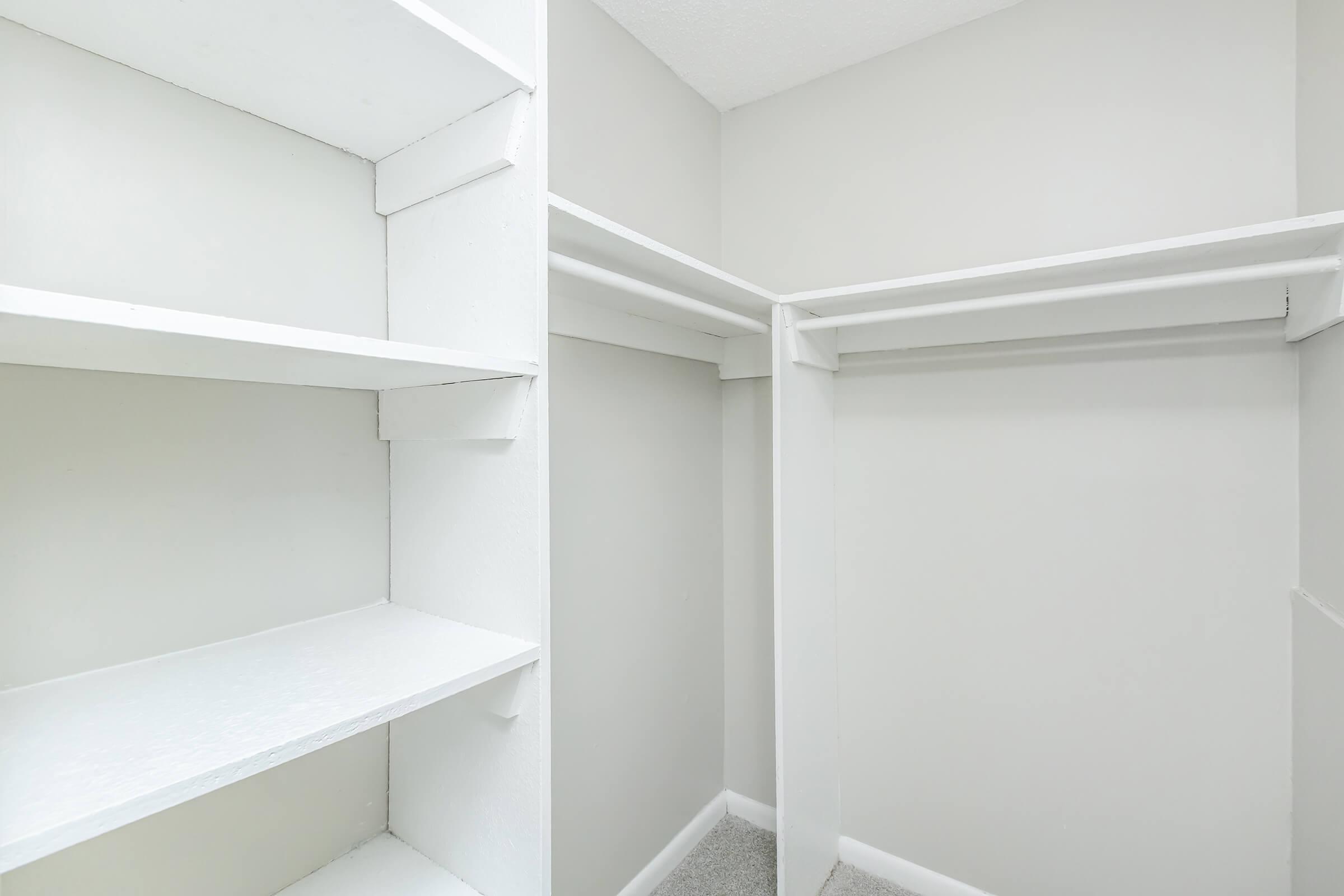 Empty closet interior featuring white shelving and hanging rods against a light gray wall. The space is well-lit and organized, with carpeted flooring.