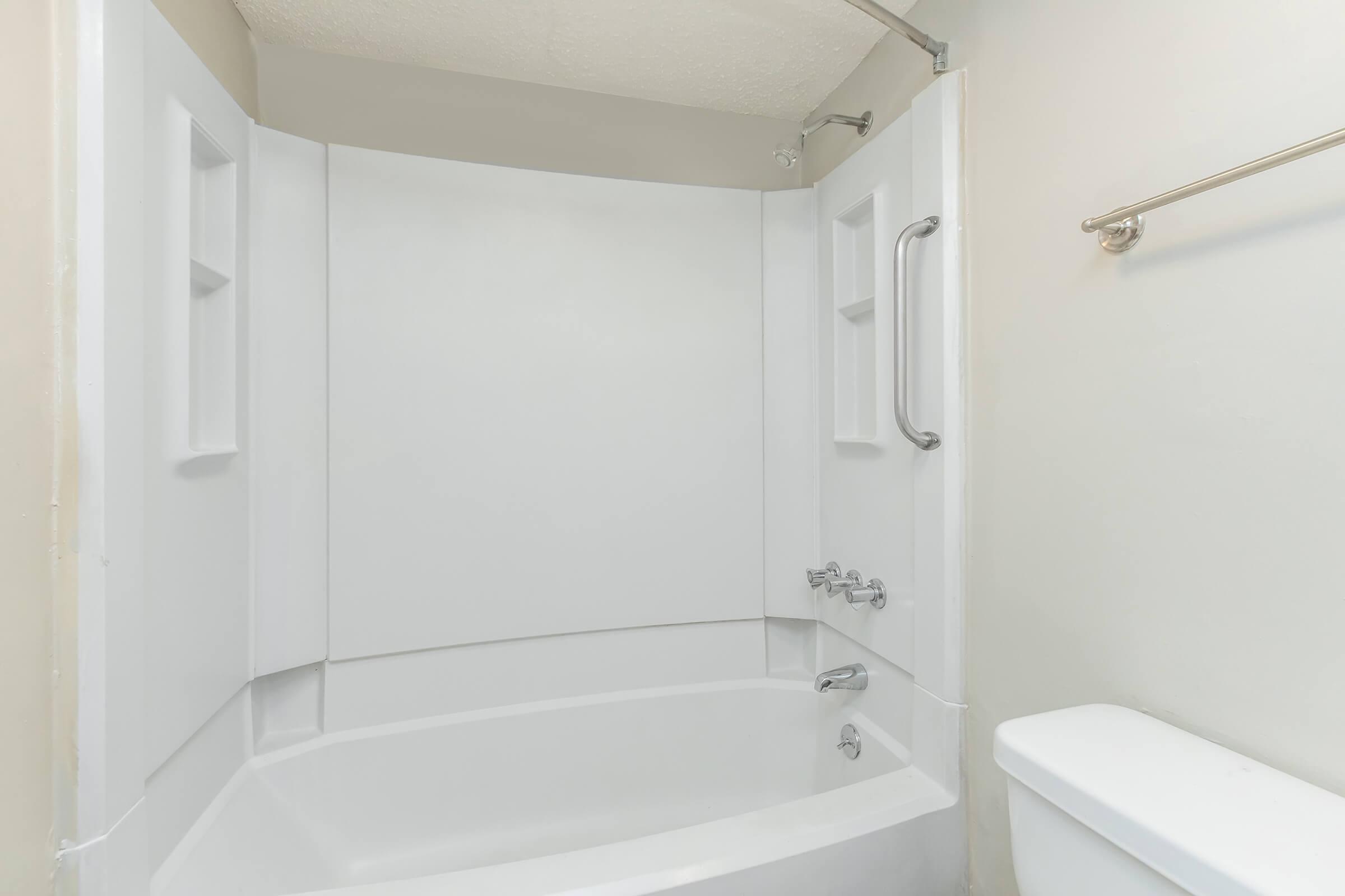 A clean, modern bathroom featuring a white bathtub with a shower curtain, a chrome faucet, and a grab bar. The walls are a neutral light color, and there is a toilet adjacent to the tub. The space is simple and uncluttered, emphasizing functionality.