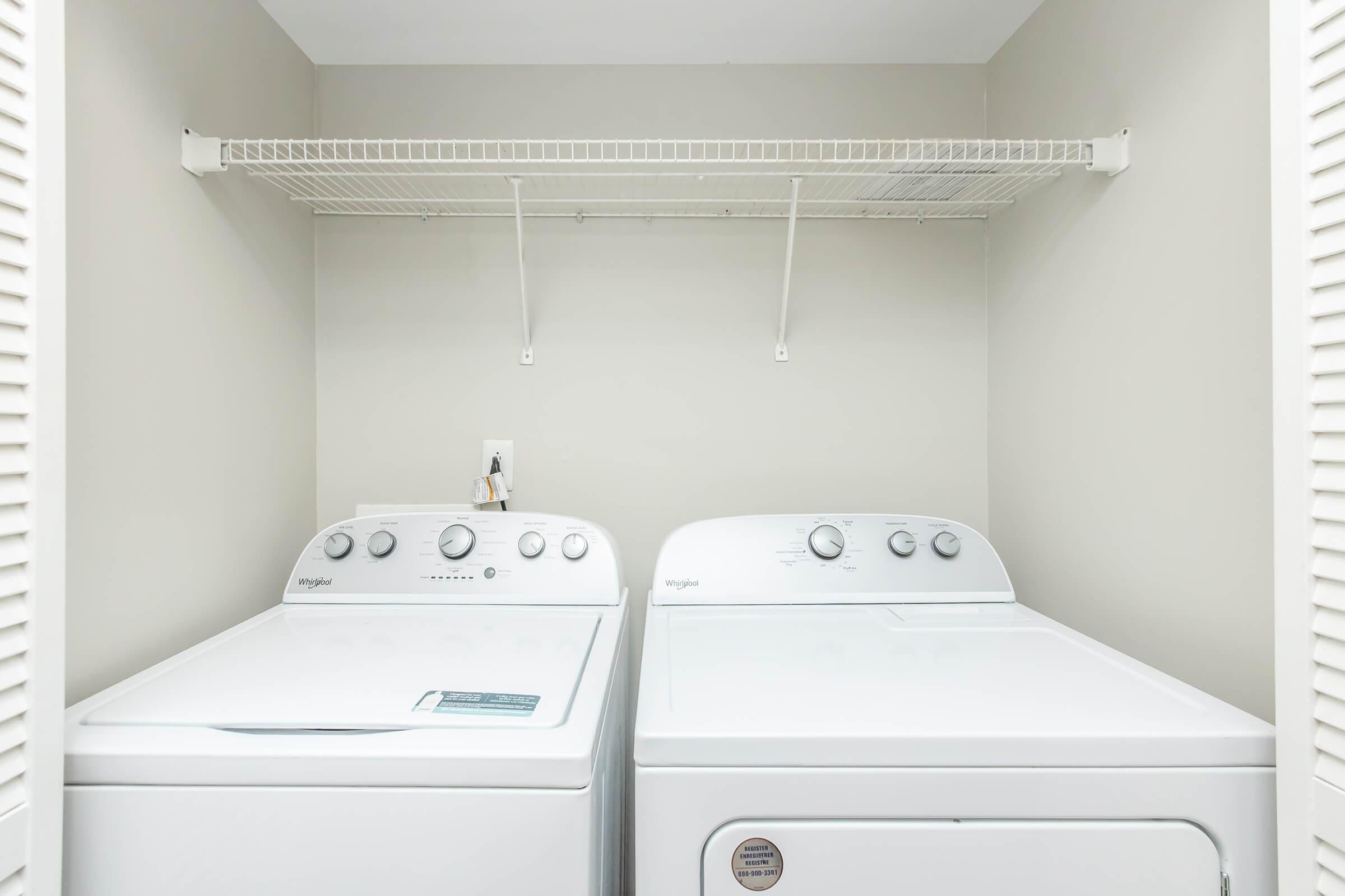 A clean laundry room featuring a white washer and dryer side by side, with a shelf above for storage. The walls are painted light gray, and there is a power outlet visible on the wall behind the appliances. The space is well-organized and tidy.