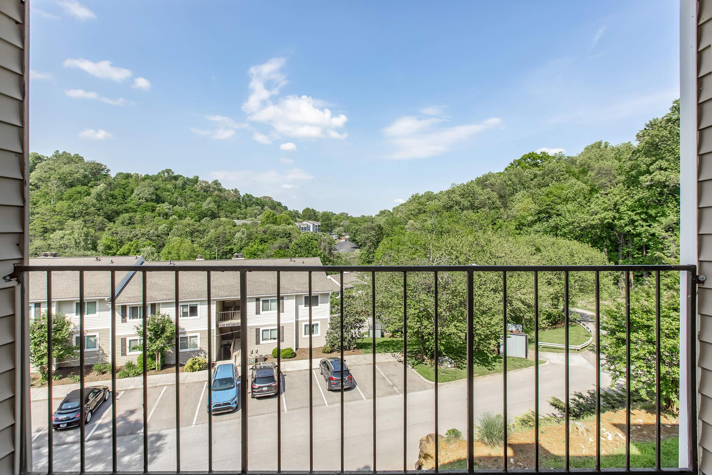 View from a balcony overlooking a green landscape and parking area. In the foreground, a railing frames the scene. Below are several parked cars and a multi-story building. The background features rolling hills and trees under a partly cloudy sky.