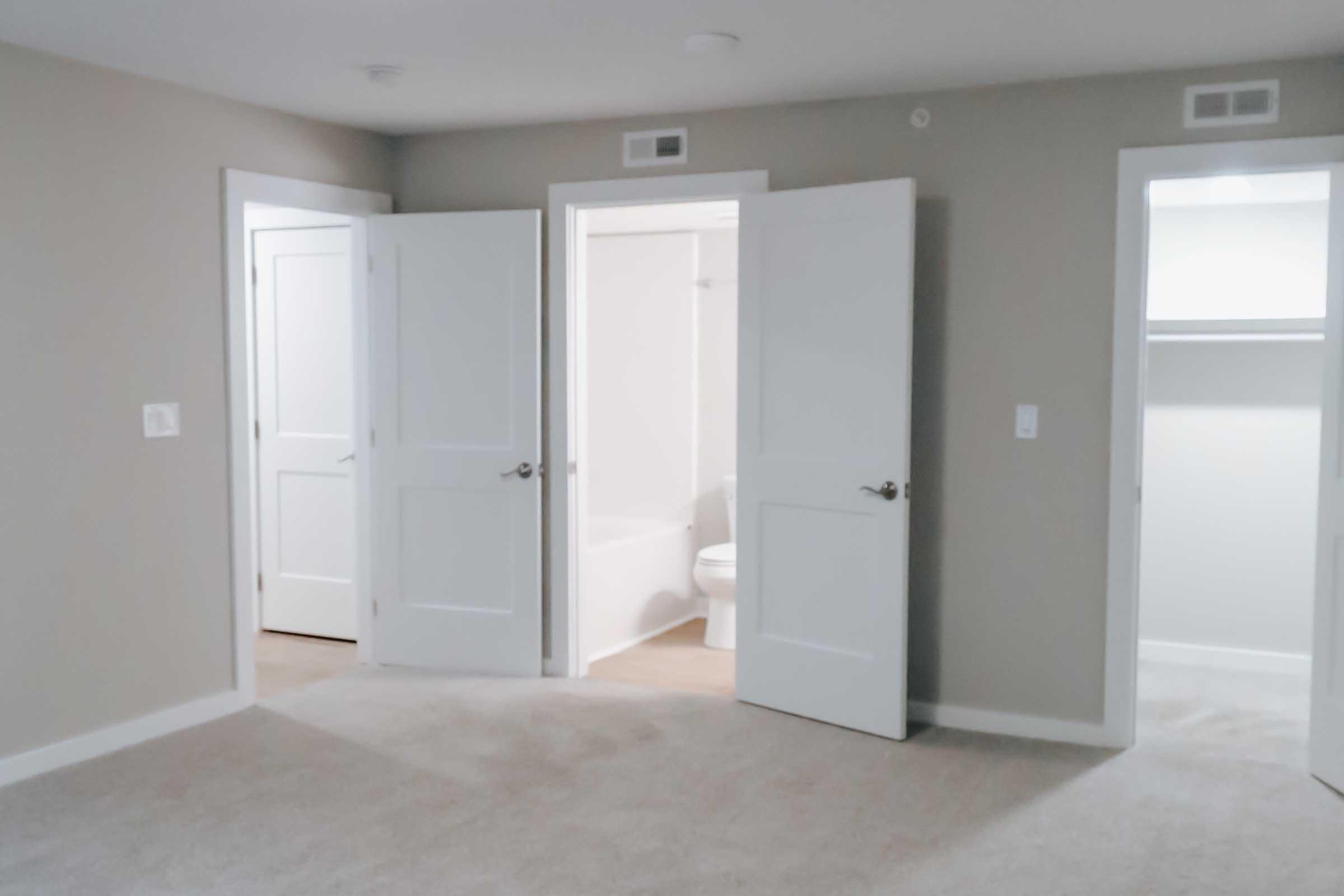 A spacious room with light-colored walls and carpet, featuring three open doors. Two doors lead to adjacent rooms, while the third door opens to a bathroom with a bathtub and toilet visible. The room is well-lit and has a neutral color scheme.