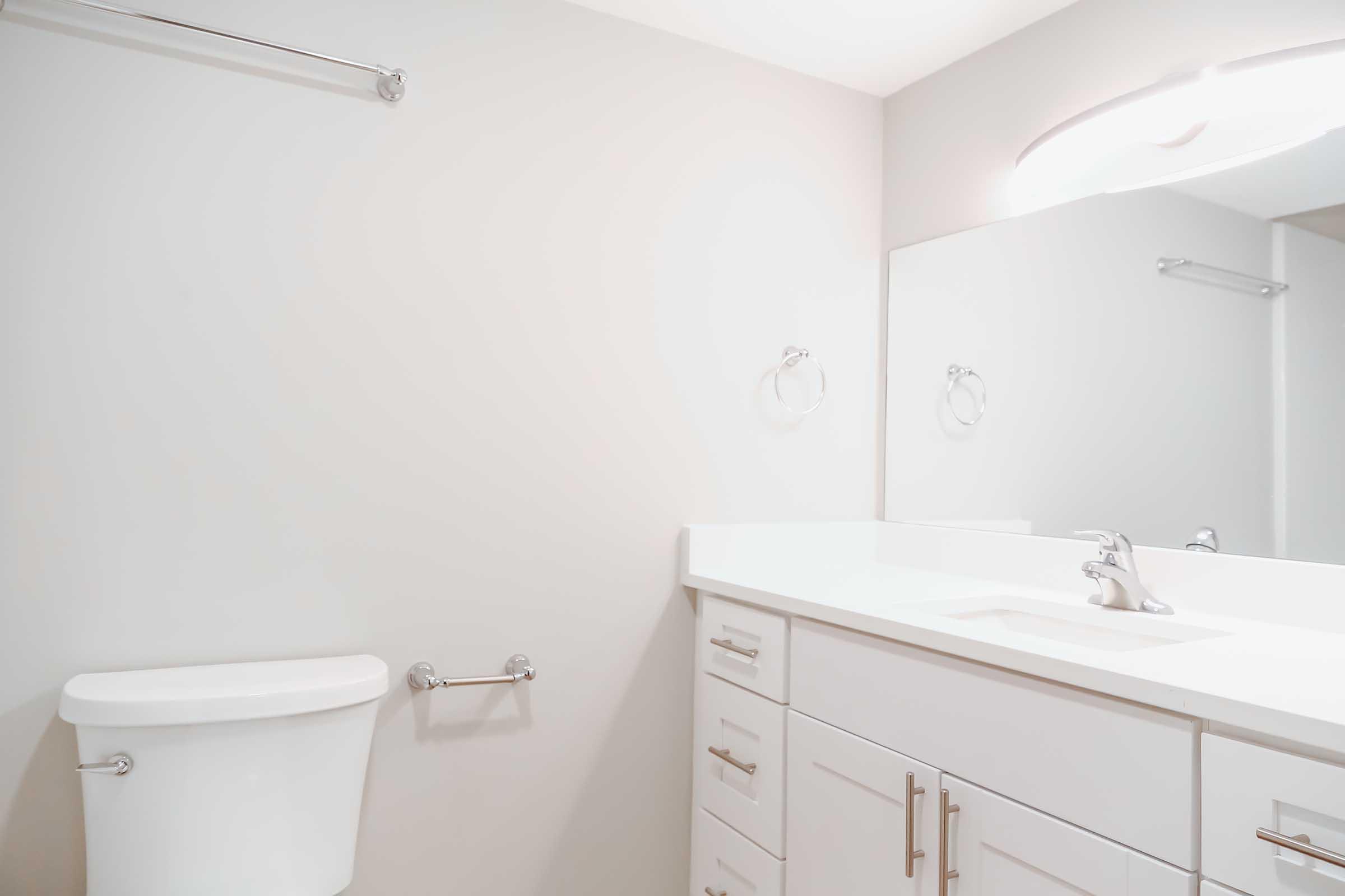 A modern bathroom featuring a white toilet, a clean white vanity with drawers, and a sink. The walls are painted light gray, and there is a mirror above the sink with a strip light. A towel bar and a hand towel holder are mounted on the walls.