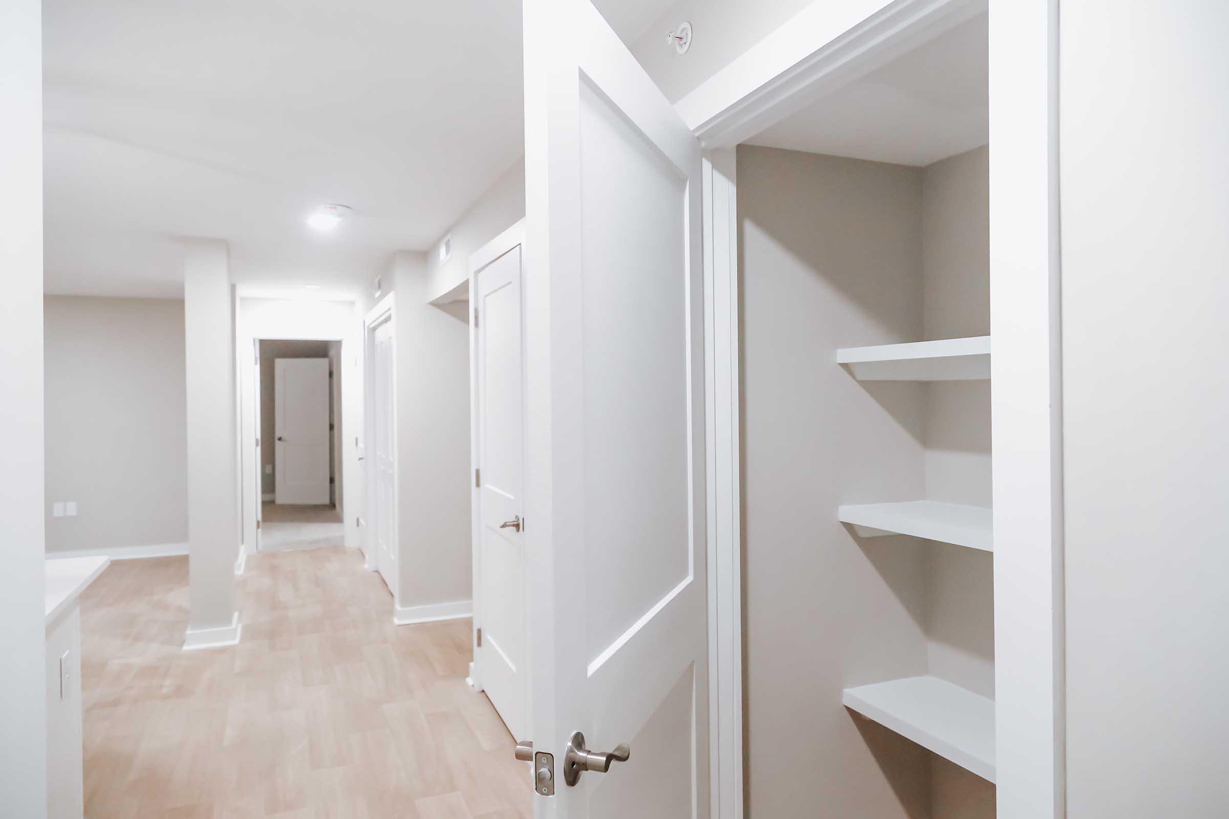 A view of a light-colored hallway with multiple doors. One door is ajar, revealing a small storage closet with shelves. The walls are painted in neutral tones, and the flooring is light wood.