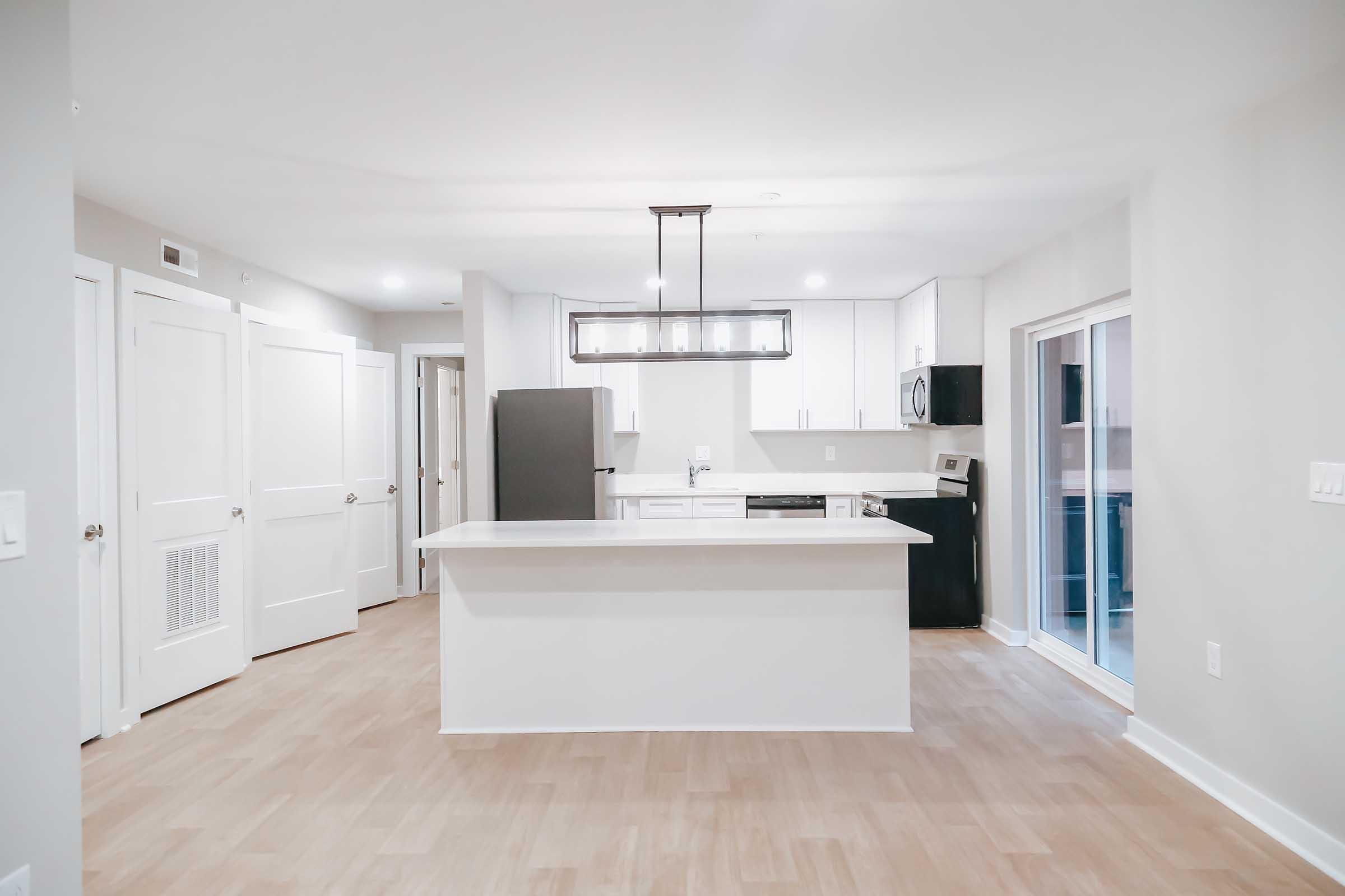 Modern kitchen interior featuring a white island with a light fixture above, stainless steel appliances, and an open layout with neutral walls and wooden flooring. The space includes a refrigerator, a stove, and sliding glass doors leading outside.