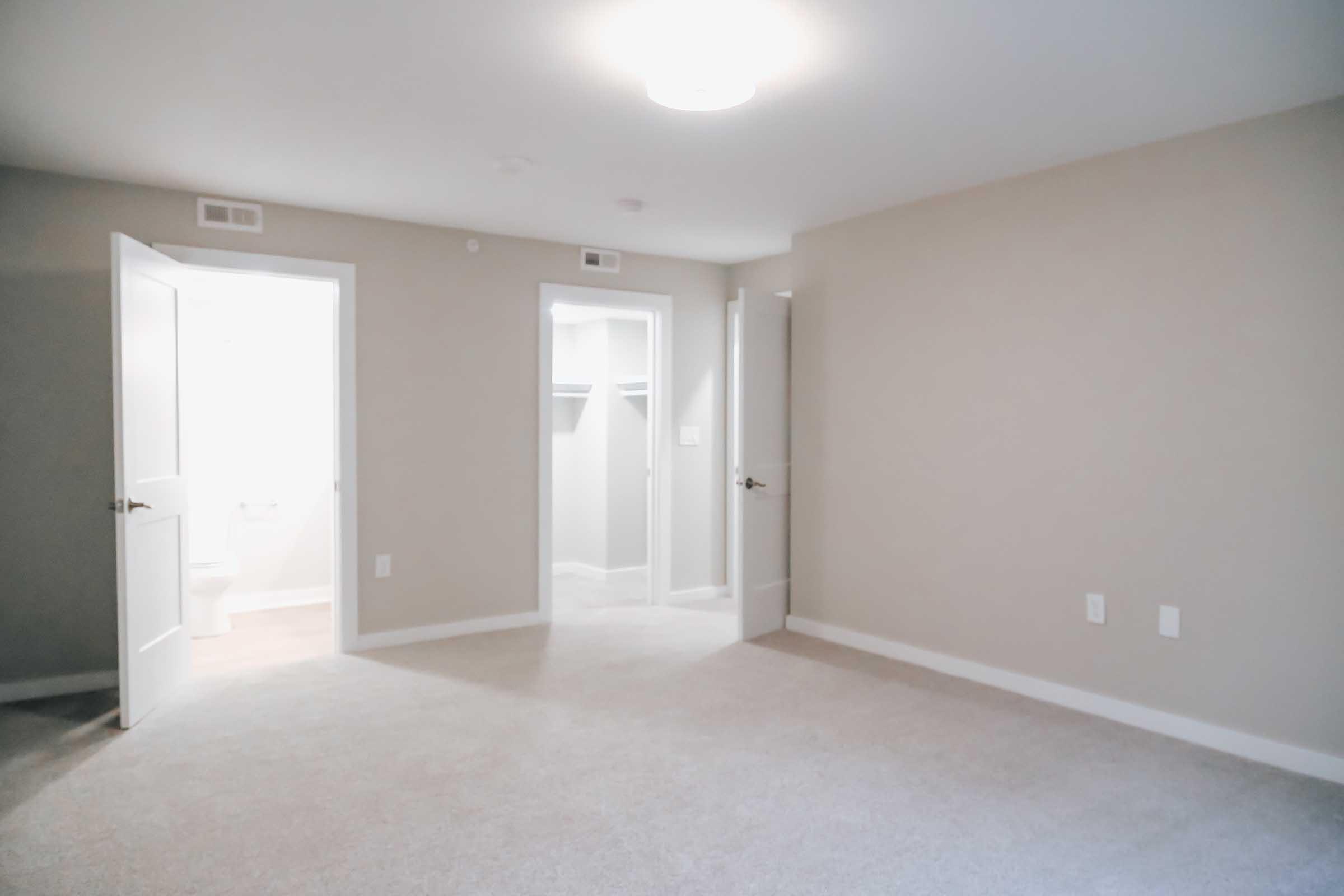 A spacious, empty room with light gray walls and carpet, featuring two open doors leading to a bathroom and a closet. The room is well-lit with a ceiling light fixture.