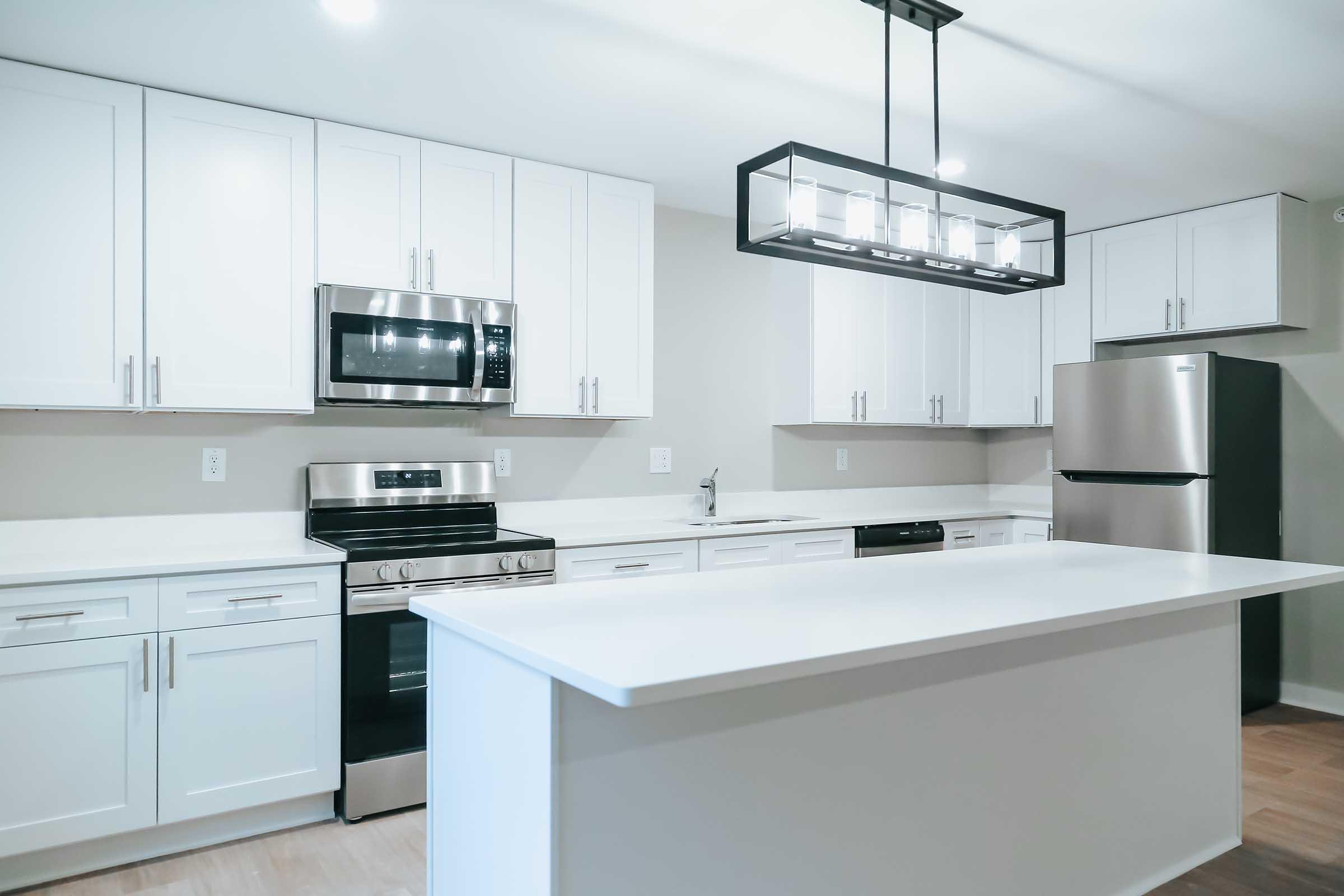 Modern kitchen featuring white cabinetry, stainless steel appliances including a microwave, oven, and refrigerator, a large island with a white countertop, and a stylish pendant light overhead.