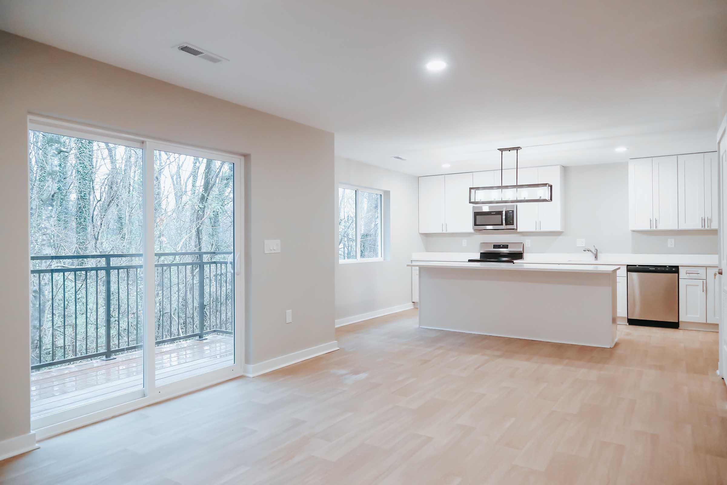 Contemporary open-concept kitchen and living area featuring a sleek design with white cabinetry, stainless steel appliances, large windows, and a sliding glass door leading to a balcony. The flooring is light-colored wood, creating a spacious and inviting atmosphere.