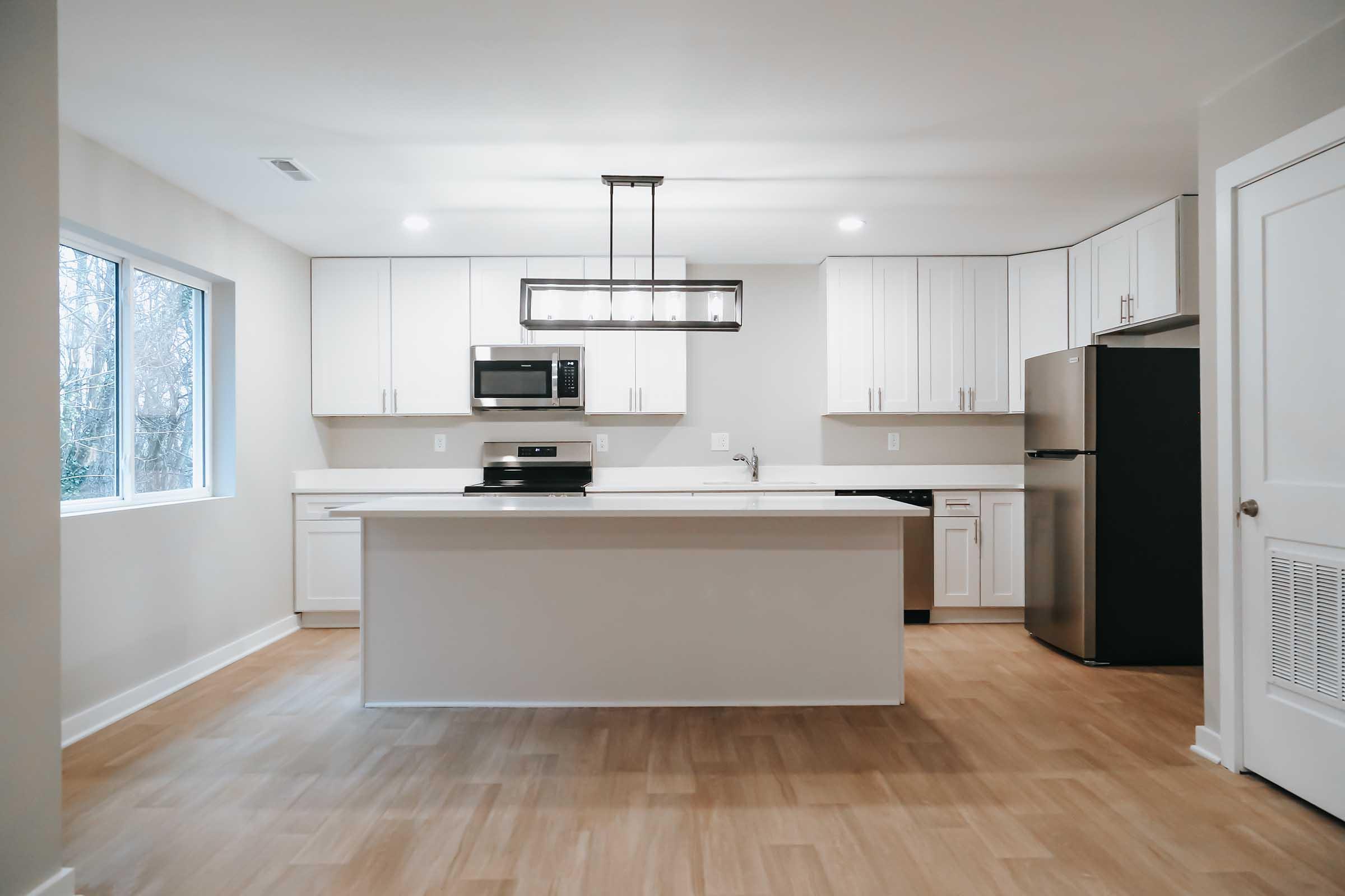 Modern kitchen interior featuring a central island, white cabinetry, stainless steel appliances including a microwave and refrigerator, and large windows providing natural light.