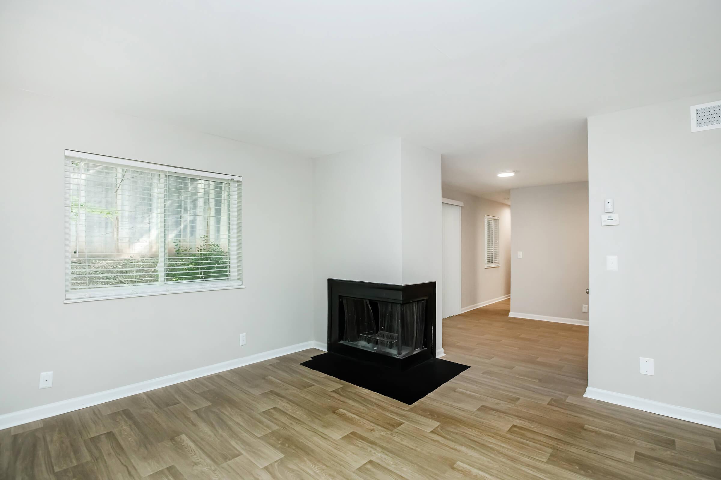 A spacious, light-filled living room with hardwood flooring, featuring a black fireplace set against a neutral wall. A window with blinds allows natural light to fill the space, while another doorway leads to a hallway. The room has a clean and modern aesthetic, perfect for relaxation or entertaining.