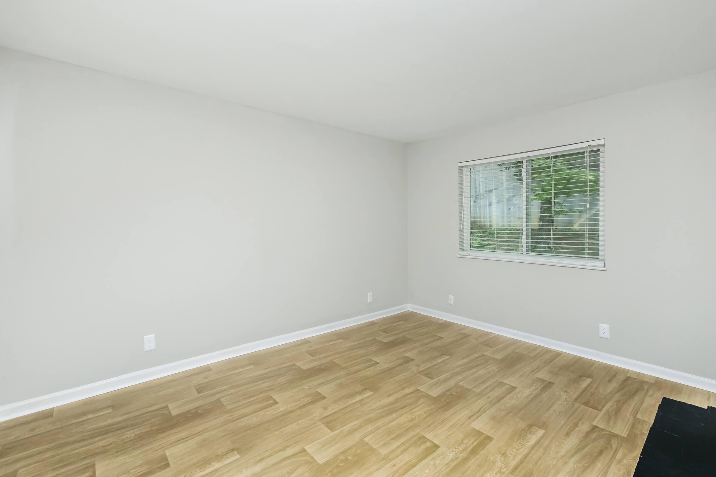 Empty room with light-colored walls and wooden flooring. A window with white blinds allows natural light into the space, and there are no furnishings present, creating a minimalist and open atmosphere.