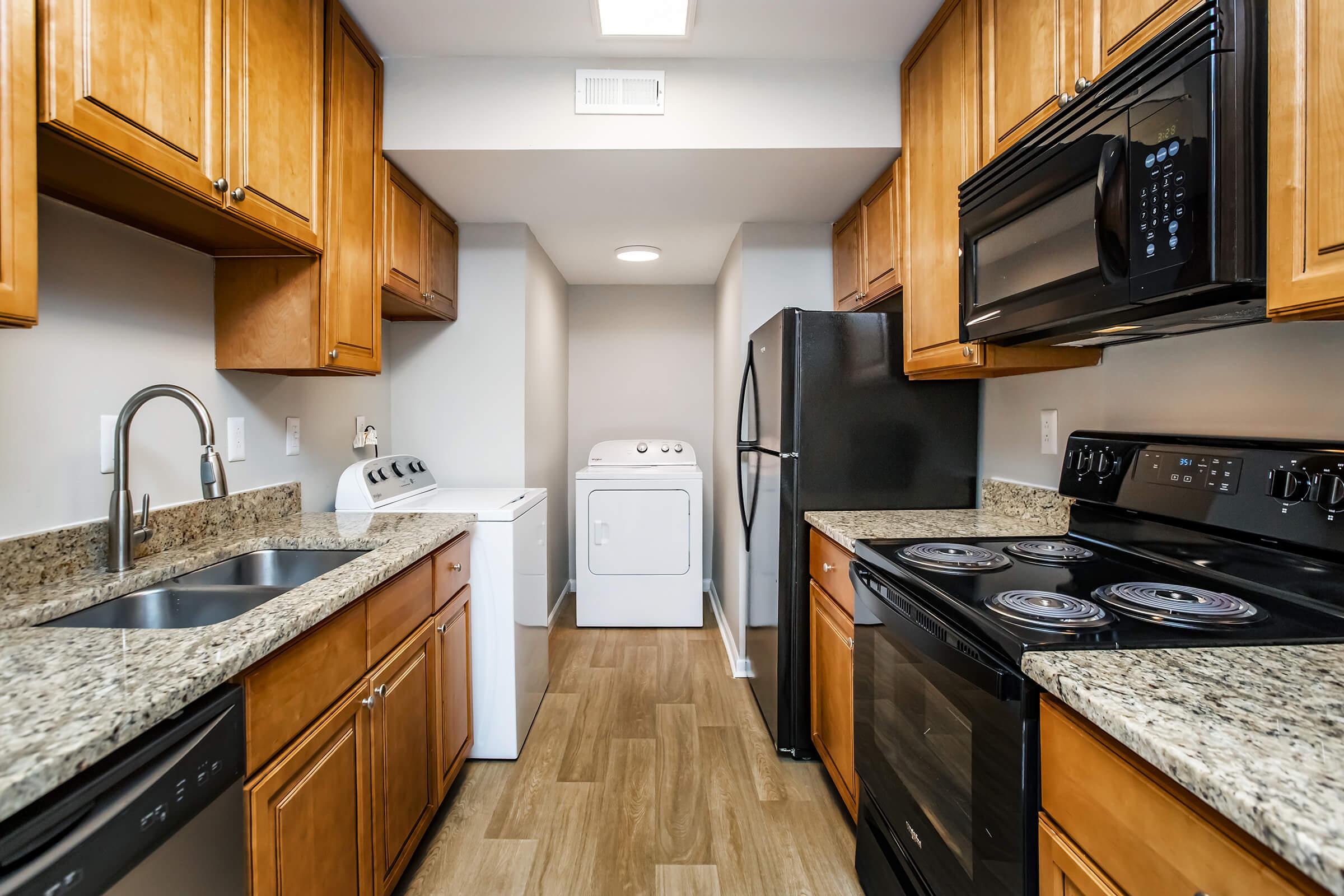 A modern kitchen with wooden cabinets and granite countertops. It features a double sink, a black refrigerator, an electric stove, and a stacked washer and dryer at the end of the room. The flooring is light-colored wood, and the walls are painted a neutral shade.