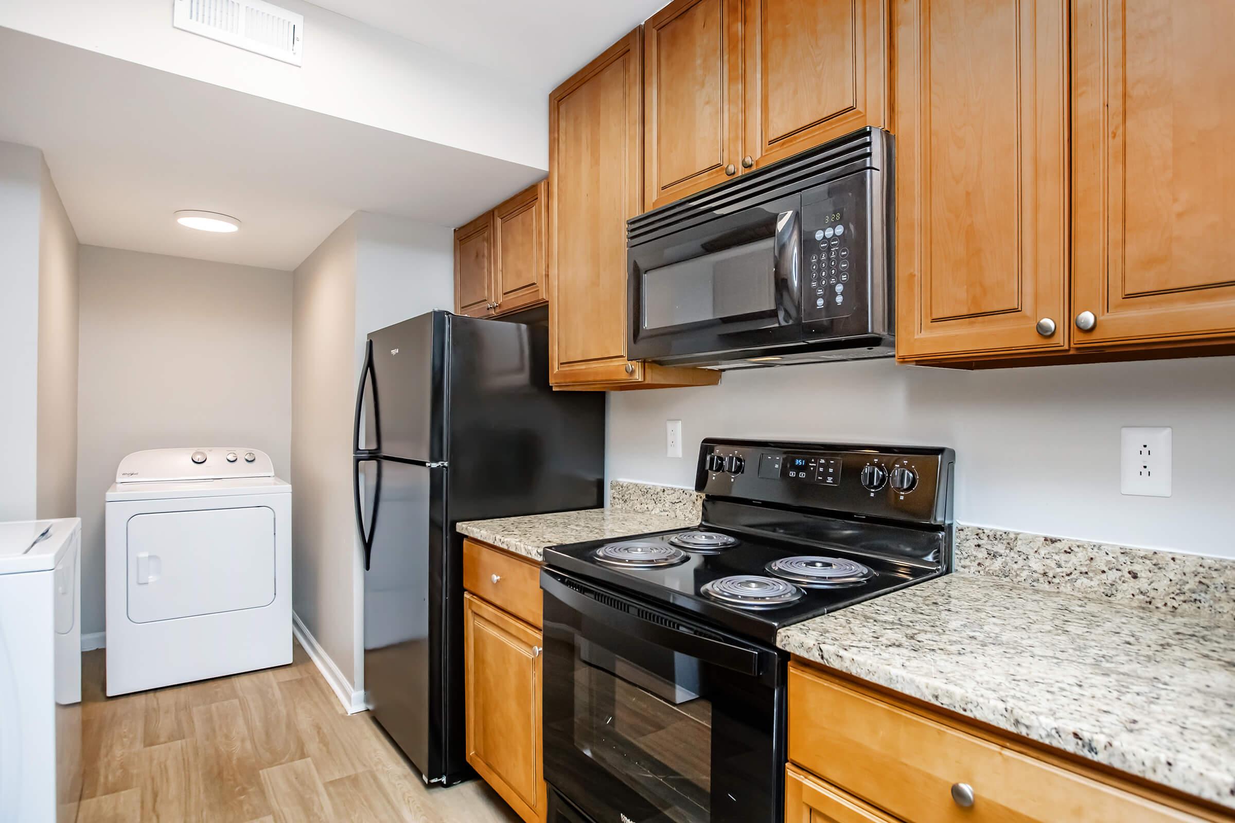 Modern kitchen with wooden cabinets and granite countertops, featuring a black stove, microwave, and refrigerator. In the background, there is a laundry area with a white washing machine and dryer. Soft lighting enhances the functional and inviting atmosphere of the space.
