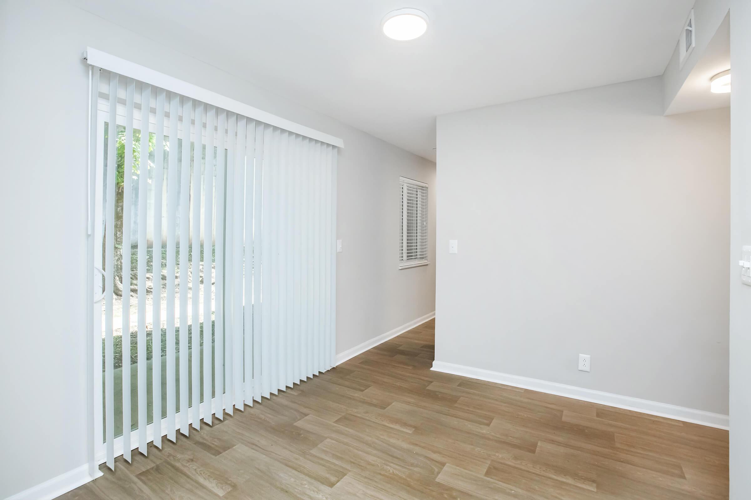 A well-lit, empty room with light-colored walls and wooden flooring. There are vertical blinds covering a sliding glass door, and a window with horizontal blinds on the left. The room appears spacious and clean, suitable for various uses.