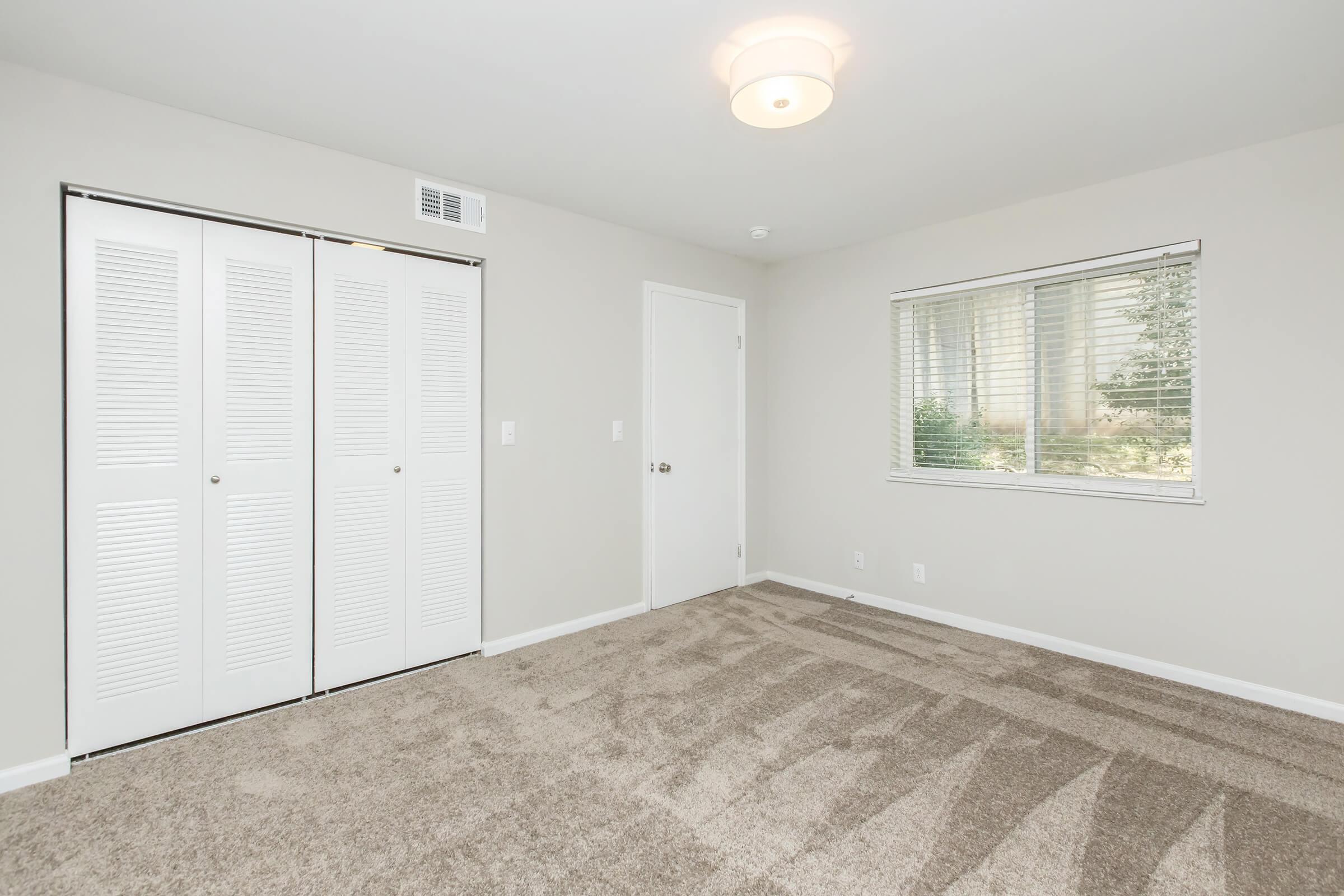 A small, well-lit bedroom featuring light grey walls, beige carpet, and a simple ceiling light fixture. There is a window on one wall with blinds and a white closet with sliding doors on the opposite wall. The room is empty, creating a clean and open space.