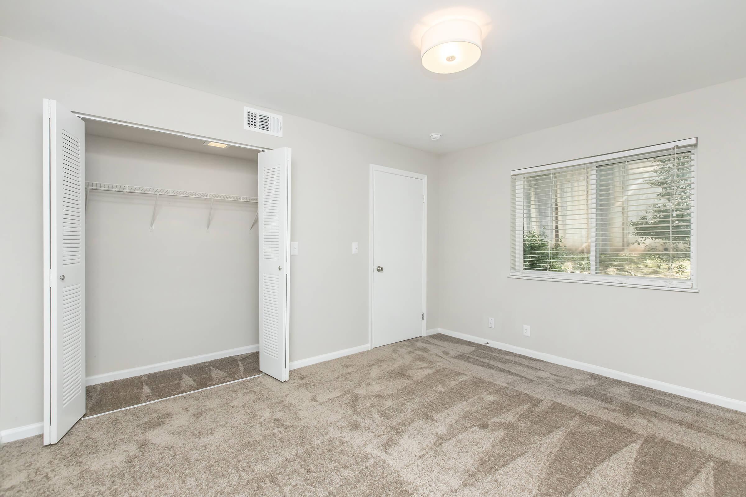 A well-lit, empty room featuring beige walls and carpet. On one side, there is a closet with sliding white doors, and a window with blinds allows natural light to enter. The door to the room is closed, and the ceiling has a simple light fixture. The overall ambiance is clean and spacious.
