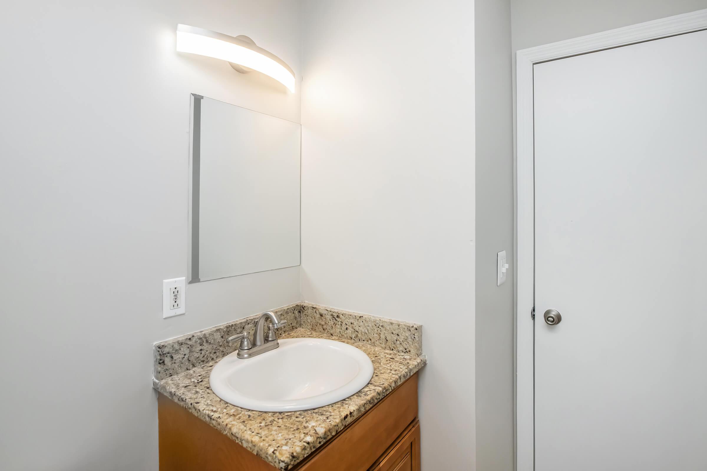 A modern bathroom setup featuring a granite countertop with a circular sink, a wall-mounted light fixture above a rectangular mirror, and a white door adjacent to the sink area. The walls are painted light gray, creating a clean and bright atmosphere.