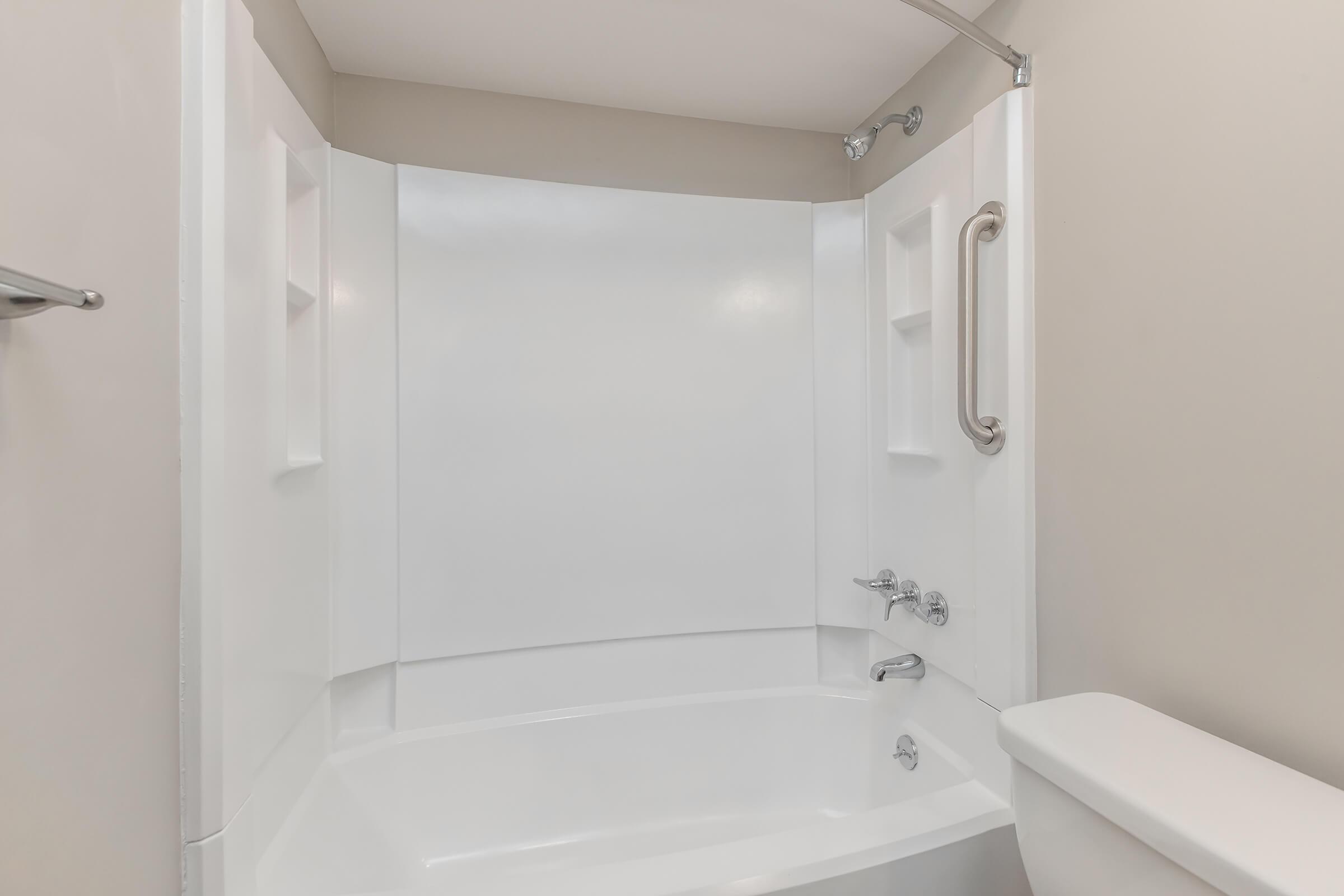 A clean, empty bathroom featuring a white bathtub with a showerhead, grab bar, and chrome fixtures. The walls are painted in a neutral color, and the space has a modern, minimalistic design, emphasizing functionality. A toilet is positioned adjacent to the bathtub.