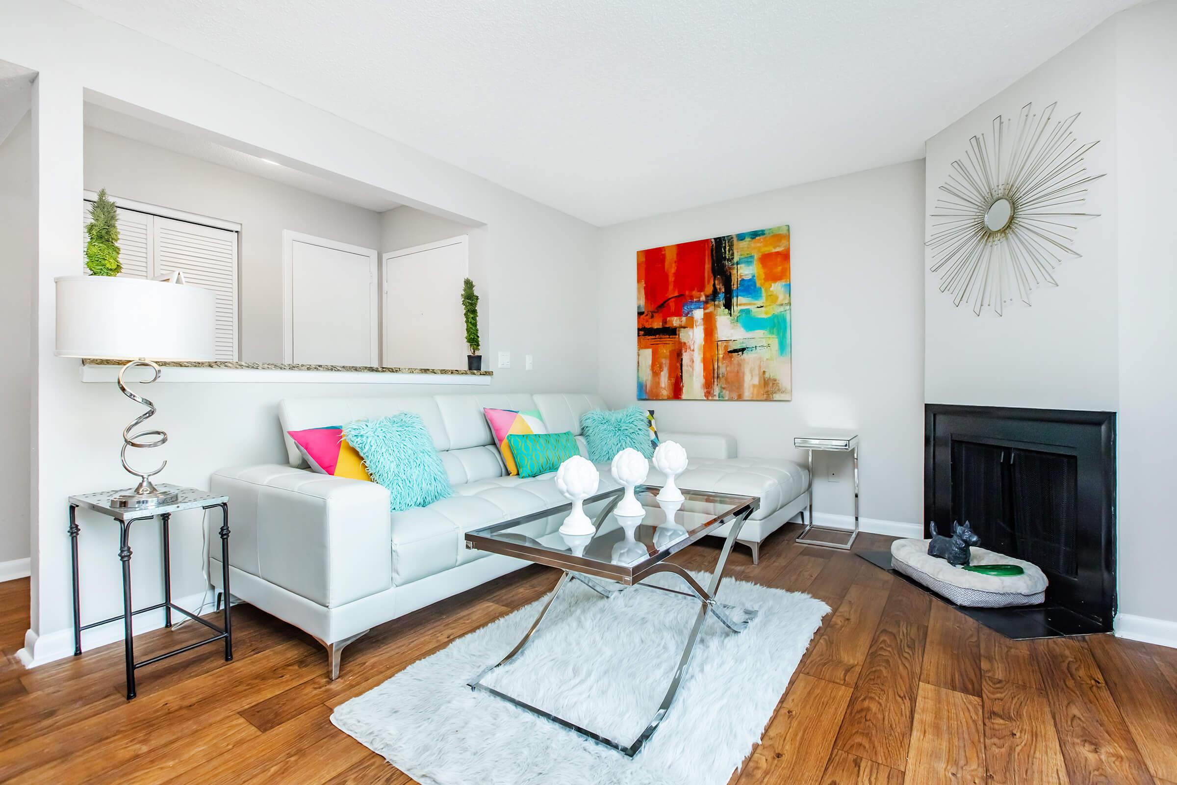 A modern living room featuring a white sectional sofa with colorful cushions, a glass coffee table on a fluffy rug, and a decorative lamp. The walls are painted in light colors, with an abstract painting and a mirror adding visual interest. A small fireplace is visible in the corner.