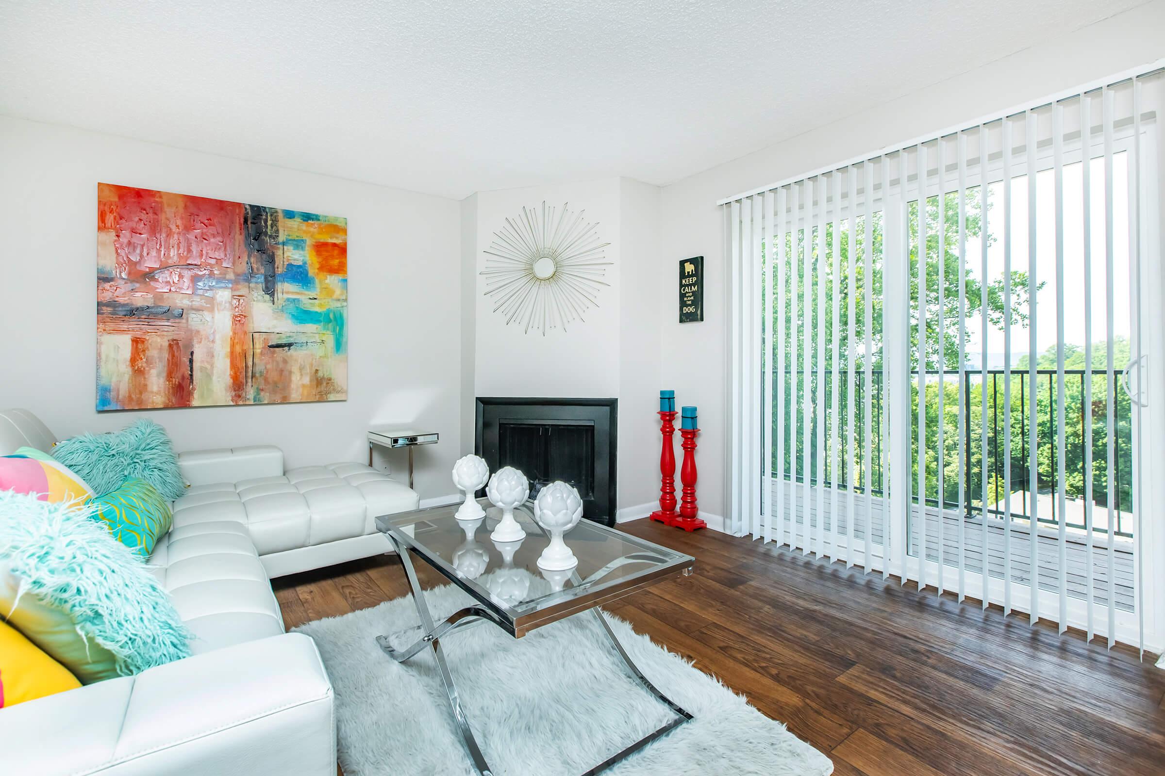 A modern living room featuring a white sectional sofa adorned with colorful pillows. There's a glass coffee table with decorative items, a vibrant abstract painting on the wall, a round mirror, and large windows with vertical blinds allowing natural light. A small fireplace and greenery visible outdoors enhance the space.