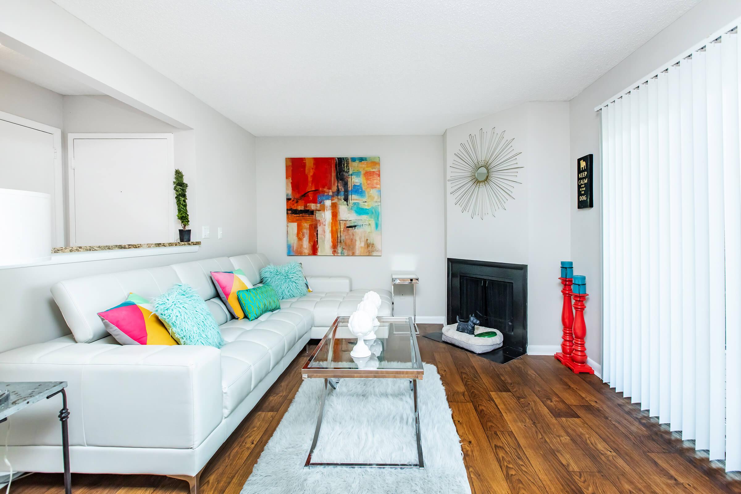 Modern living room featuring a white leather sectional sofa adorned with colorful pillows, a glass coffee table on a fluffy rug, hardwood flooring, and a decorative wall art piece. A fireplace is present, along with a sunburst mirror and accents of vibrant decor on the walls and shelves.