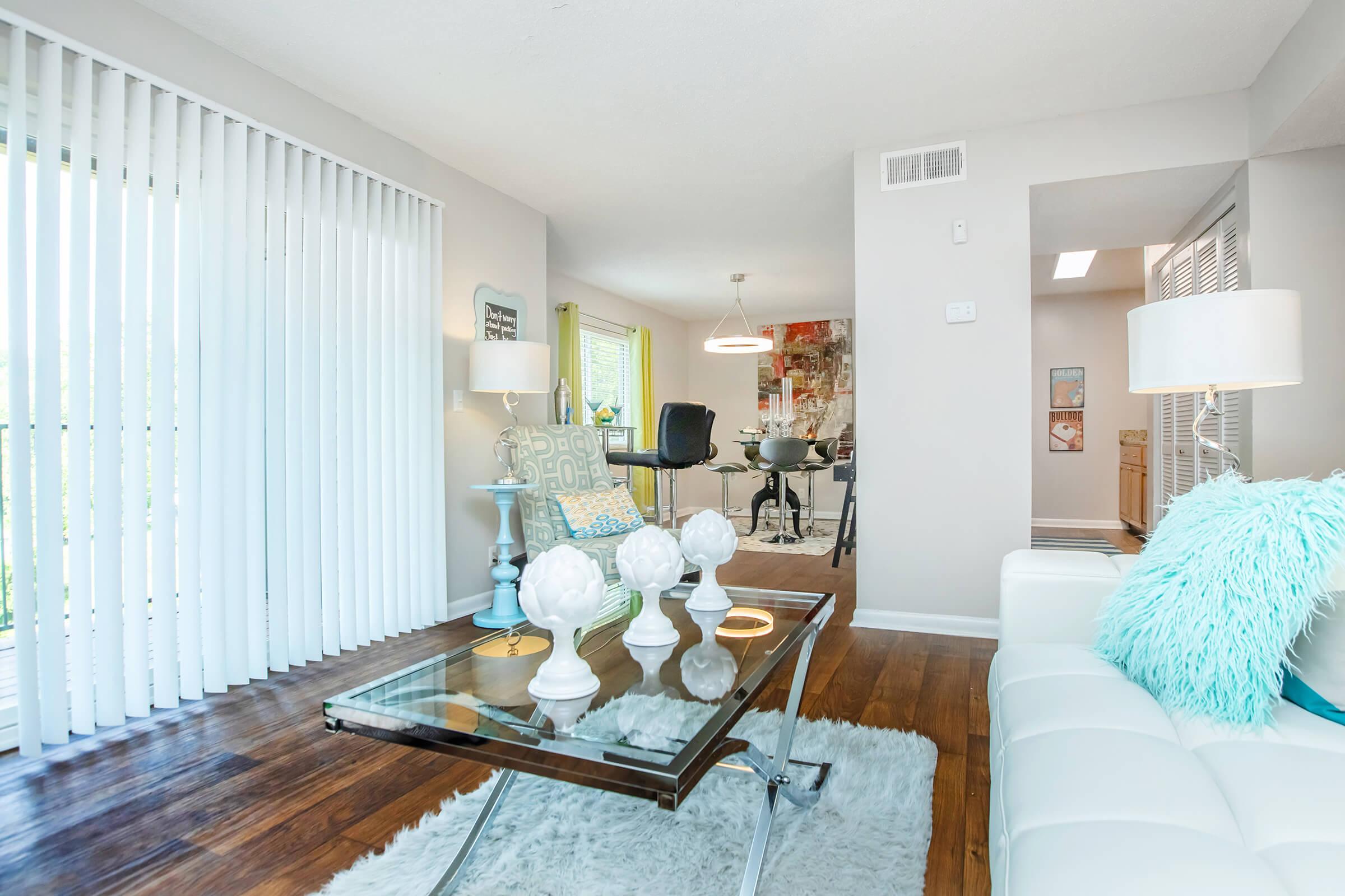 Modern living room featuring a white sofa with a blue accent pillow, a glass coffee table with decorative white ornaments, and a light-colored rug. The space includes large sliding glass doors with vertical blinds, a dining area visible in the background, and stylish lighting.