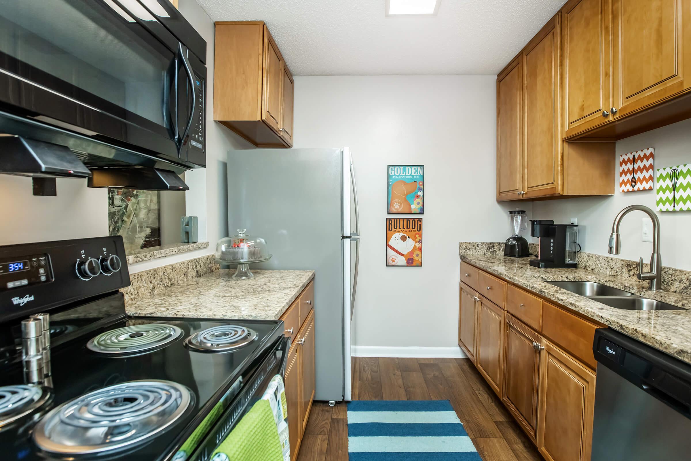 A modern kitchen featuring wooden cabinets, granite countertops, a black stove, and a stainless steel refrigerator. The space includes a coffee maker, a sink, and colorful wall art. A blue striped rug is on the floor, enhancing the contemporary design.