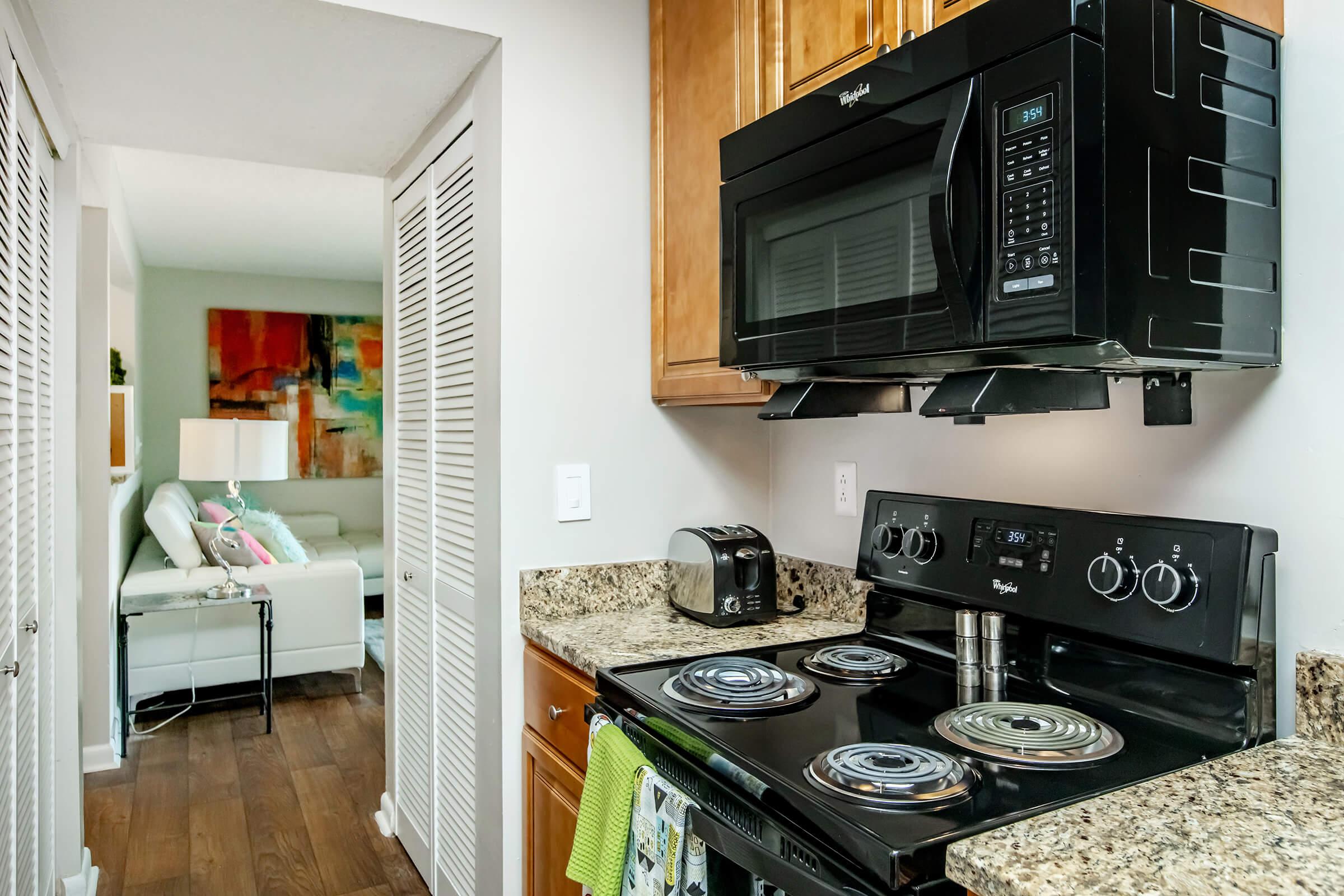 A modern kitchen featuring a black microwave above a black stove with knobs, granite countertops, and a toaster. The background includes a hallway leading to a living area with a light-colored sofa and vibrant wall art.