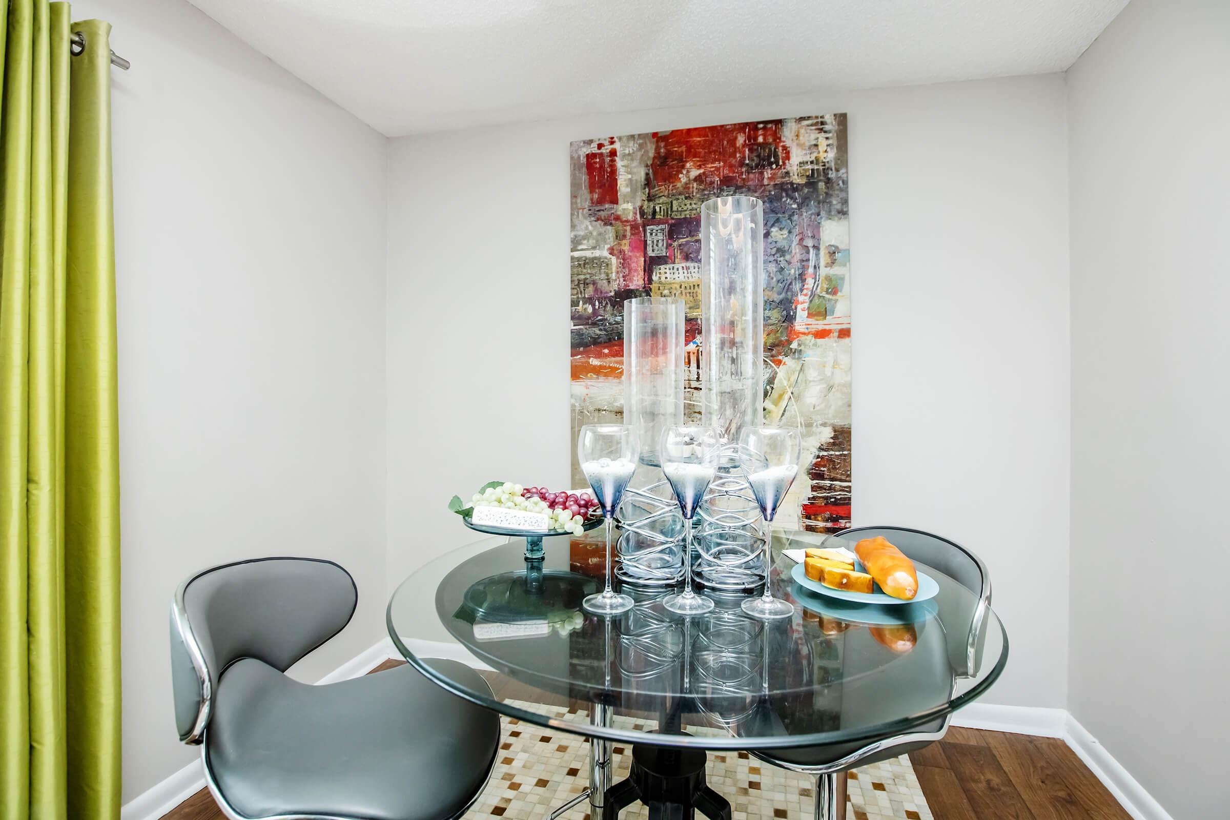 A modern dining area featuring a glass round table with elegant tall candle holders and a fruit platter. The table is surrounded by sleek chairs and placed against a colorful abstract painting on the wall. Green curtains frame the window, adding a touch of color to the neutral room.