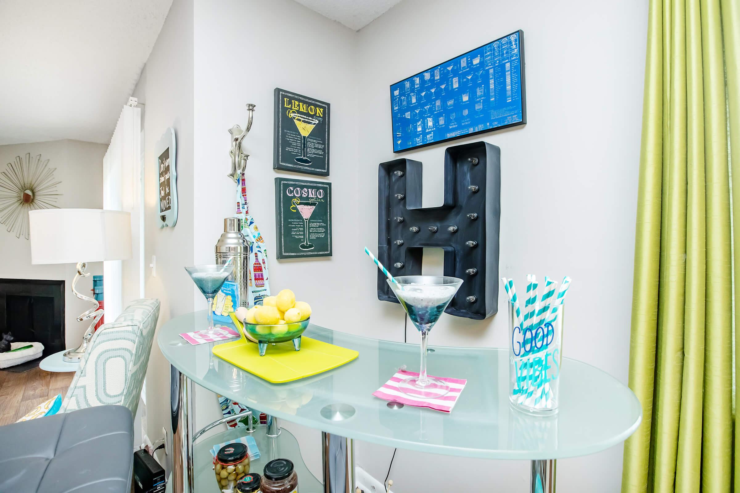 A stylish home bar area featuring a glass table with cocktails, a bowl of lemons, and colorful straws. Wall decor includes framed drink menus and a large letter "H" display. Bright green curtains add a pop of color to the modern setting, creating a cheerful atmosphere for entertaining.
