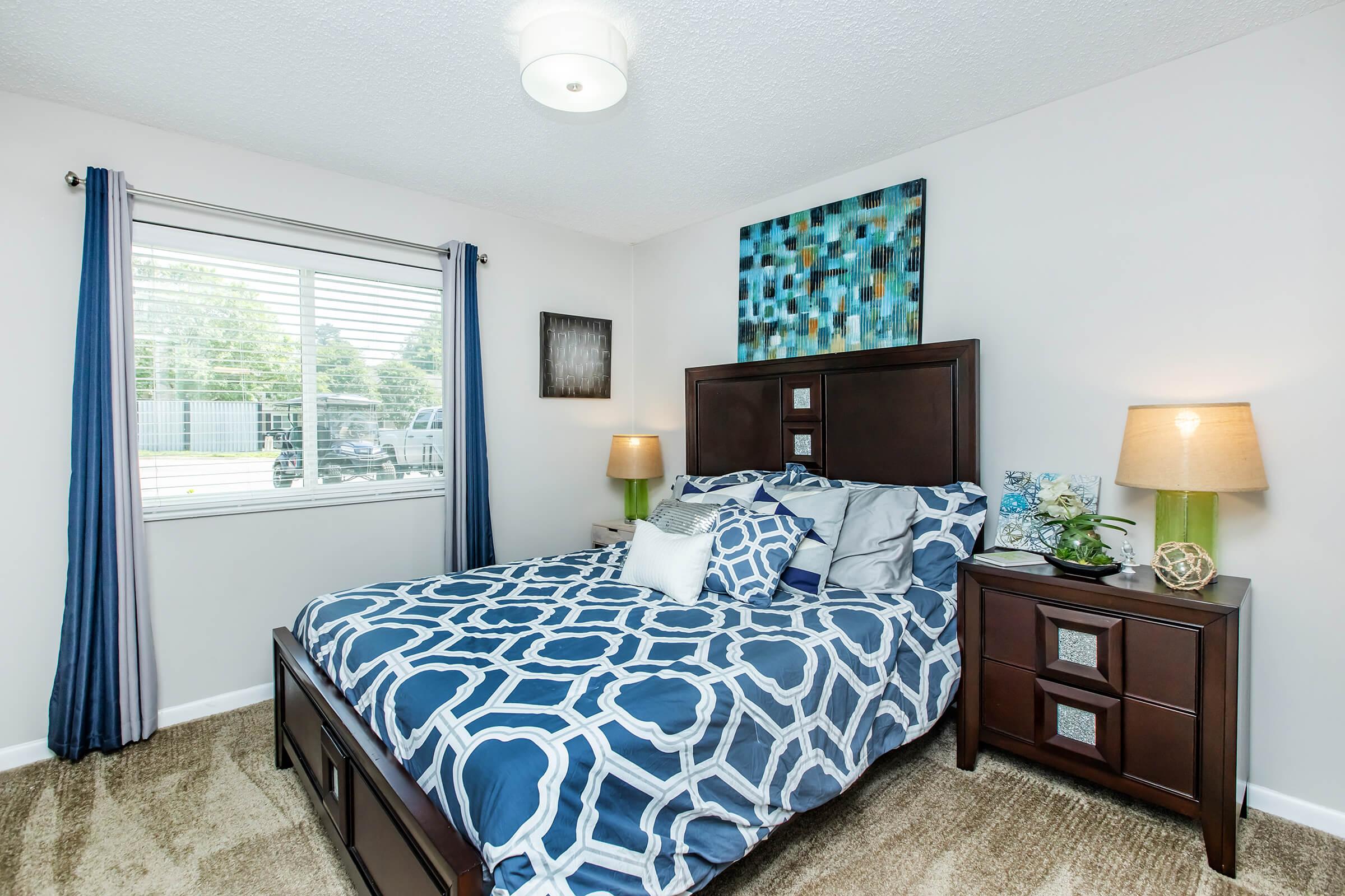 A cozy bedroom featuring a dark wooden bed with a patterned blue and white duvet, flanked by matching bedside tables with lamps. Windows with blue curtains allow natural light, and a piece of abstract artwork hangs above the bed. The room has beige carpet and a clean, inviting atmosphere.