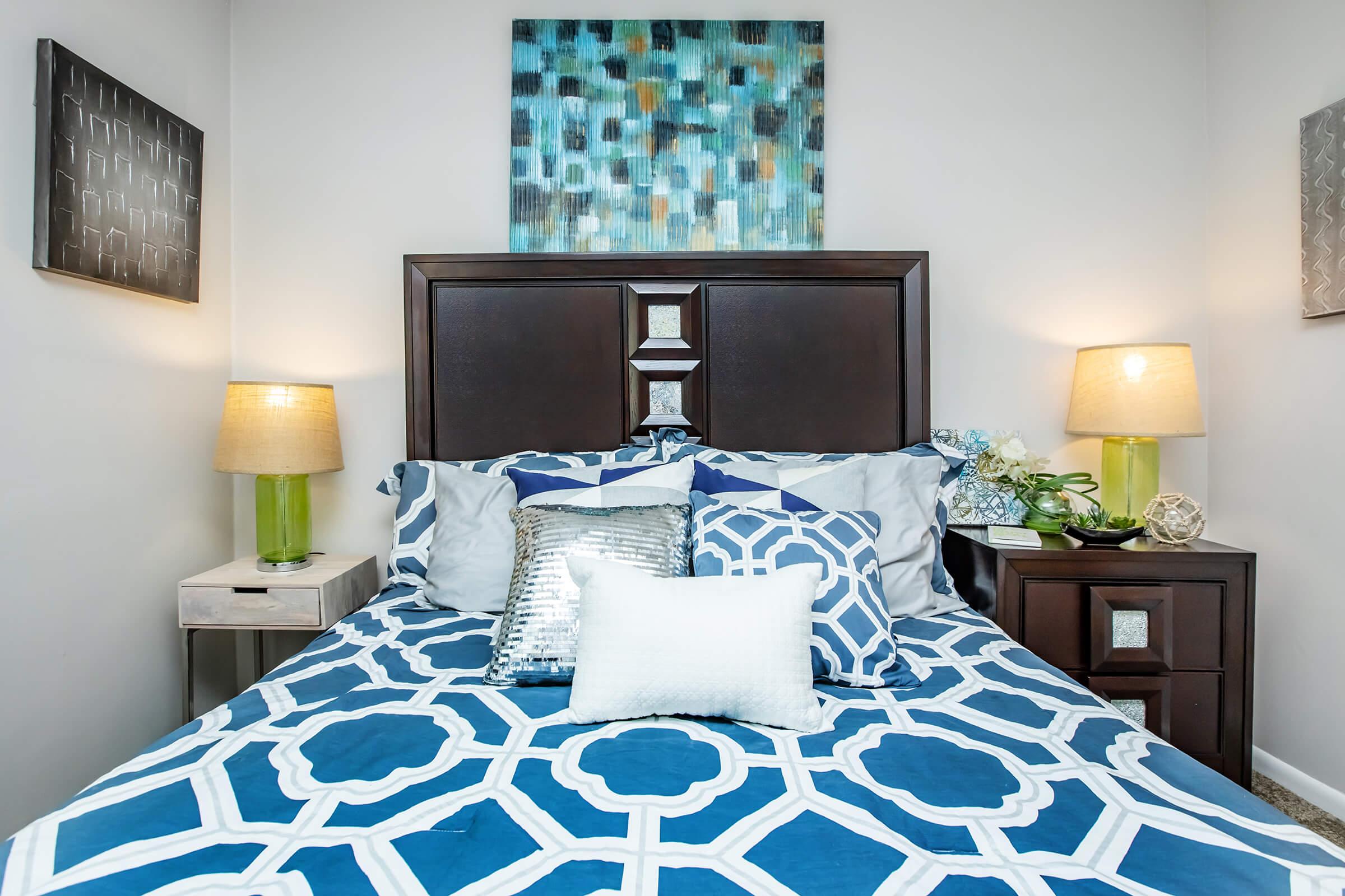 A cozy bedroom featuring a dark wood headboard with decorative details. The bed is dressed in a blue and white patterned duvet and multiple pillows. On either side of the bed are bedside tables with lamps and a small plant. Artwork hangs above the bed, adding color to the room's light, welcoming atmosphere.