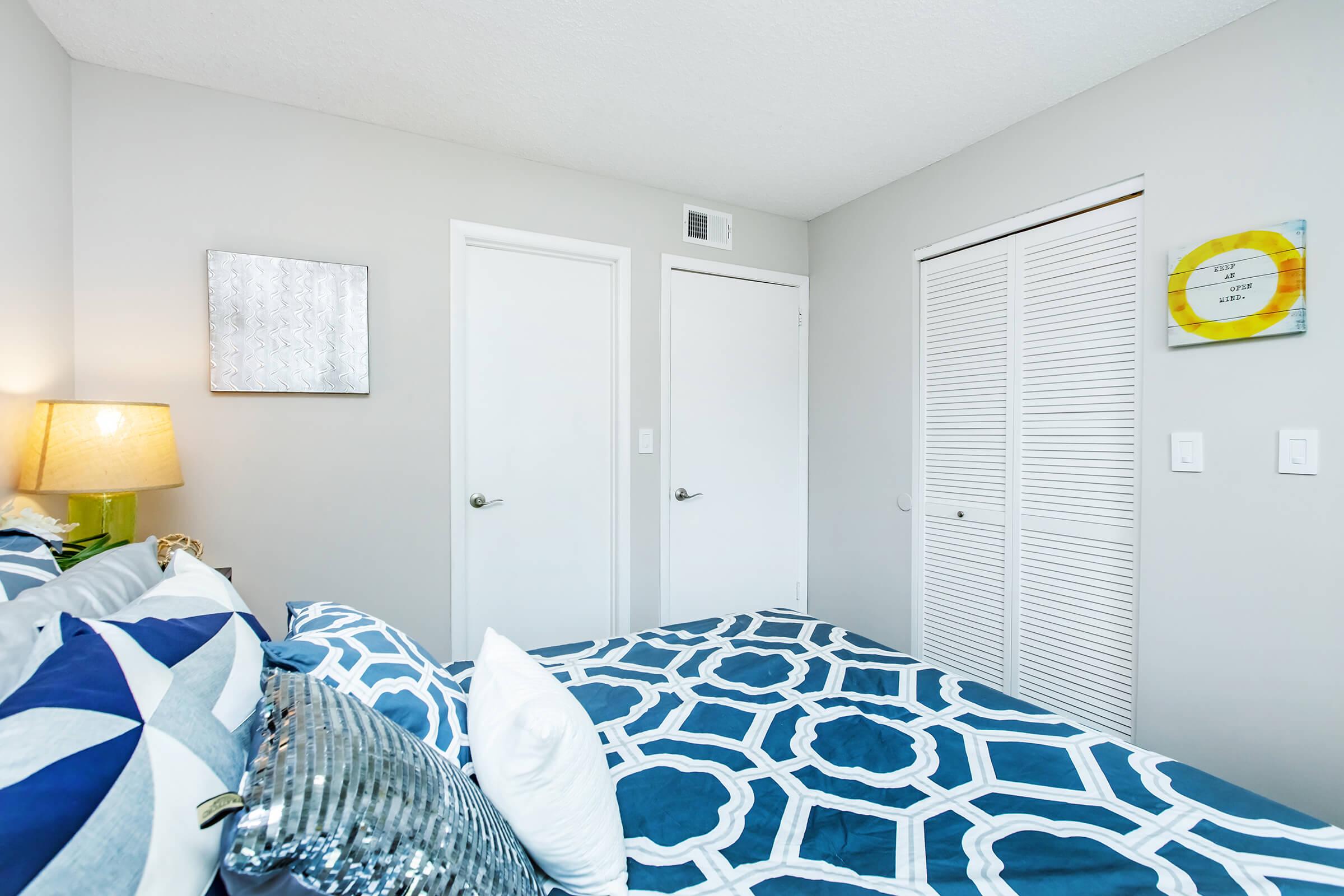 A cozy bedroom featuring a neatly made bed with patterned blue and white bedding. There are two closed doors on the back wall, a small decorative mirror, and a wall clock. Soft lighting from a bedside lamp adds warmth to the space. The walls are painted in a neutral shade, creating a calm atmosphere.