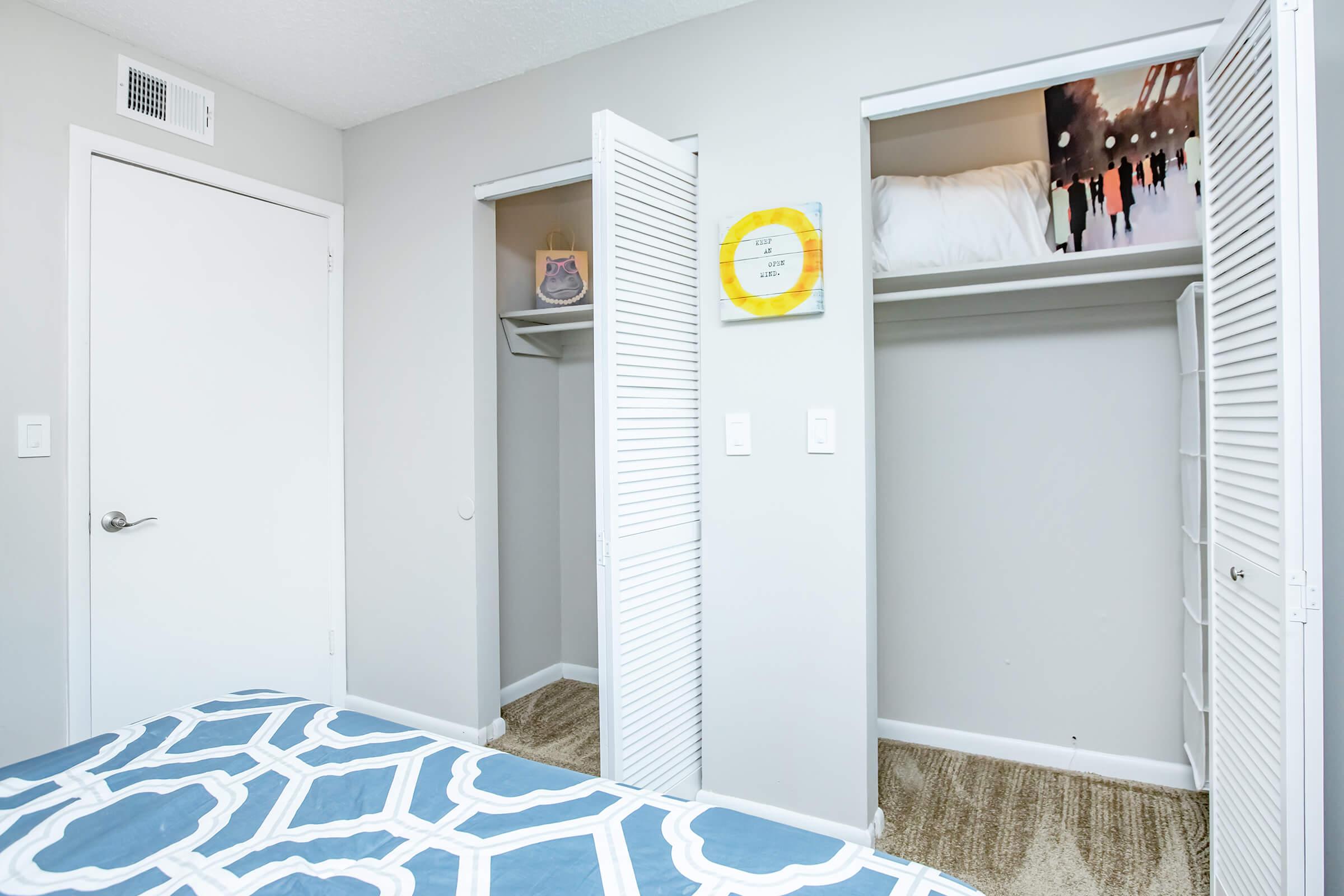 A well-lit interior view of a bedroom featuring a bed with a blue and white patterned comforter. Two closets with white louvered doors are visible, one partially open revealing shelves. The room has light gray walls and a door leading outside the frame.