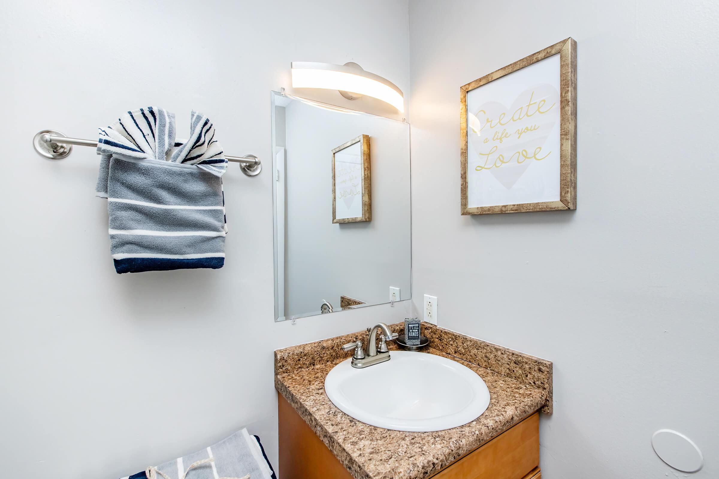 A modern bathroom featuring a round sink with a granite countertop, a wall-mounted mirror, and a framed inspirational quote. A towel rack holds neatly folded towels in blue and gray. The walls are painted light gray, and there’s a small soap dispenser near the sink.