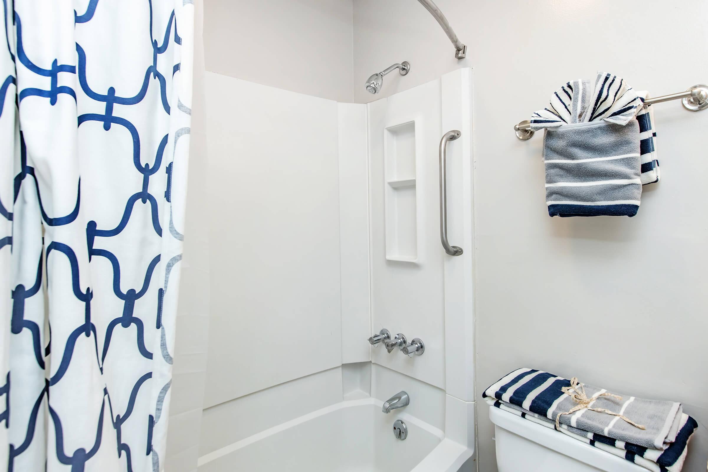 A clean, modern bathroom featuring a white bathtub, a gray and blue patterned shower curtain, and neatly arranged towels on a rack. The walls are painted light gray, and there is a built-in shelf for toiletries next to the tub, creating a functional and stylish space.