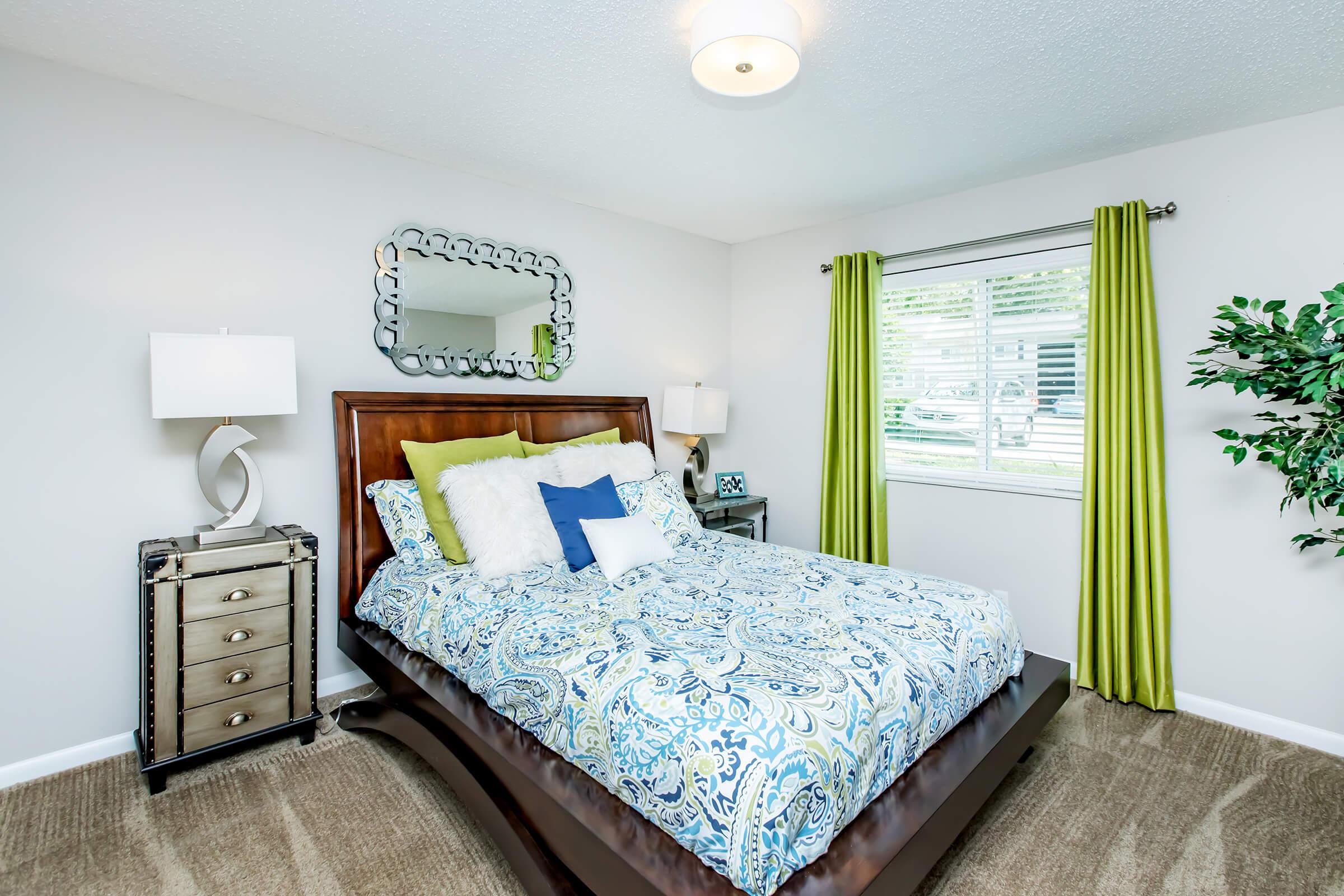 A cozy bedroom featuring a wooden bed with a patterned blue and white comforter, several decorative pillows, and a stylish headboard. There are two bedside lamps, a mirrored dresser, and large green curtains by the window. A decorative plant adds a touch of nature to the room.