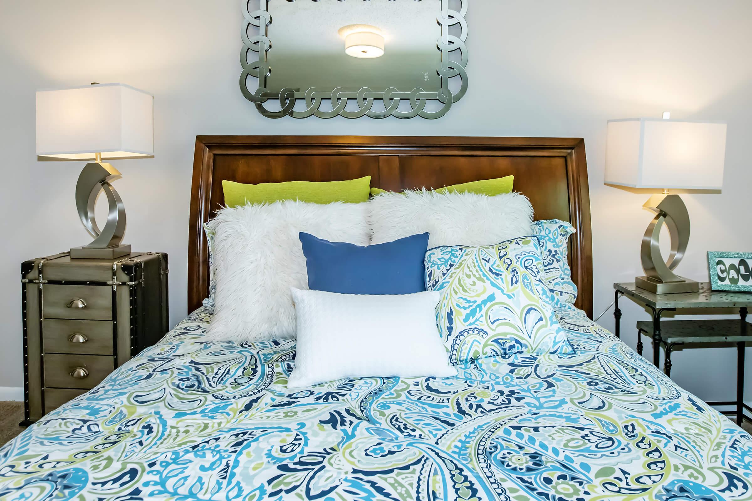 A well-decorated bedroom featuring a wooden headboard with multiple colorful decorative pillows on the bed. Two stylish lamps with curved bases flank the bed, and a decorative mirror hangs above the headboard. The bedding features a vibrant paisley pattern in shades of blue and green, creating a cozy atmosphere.