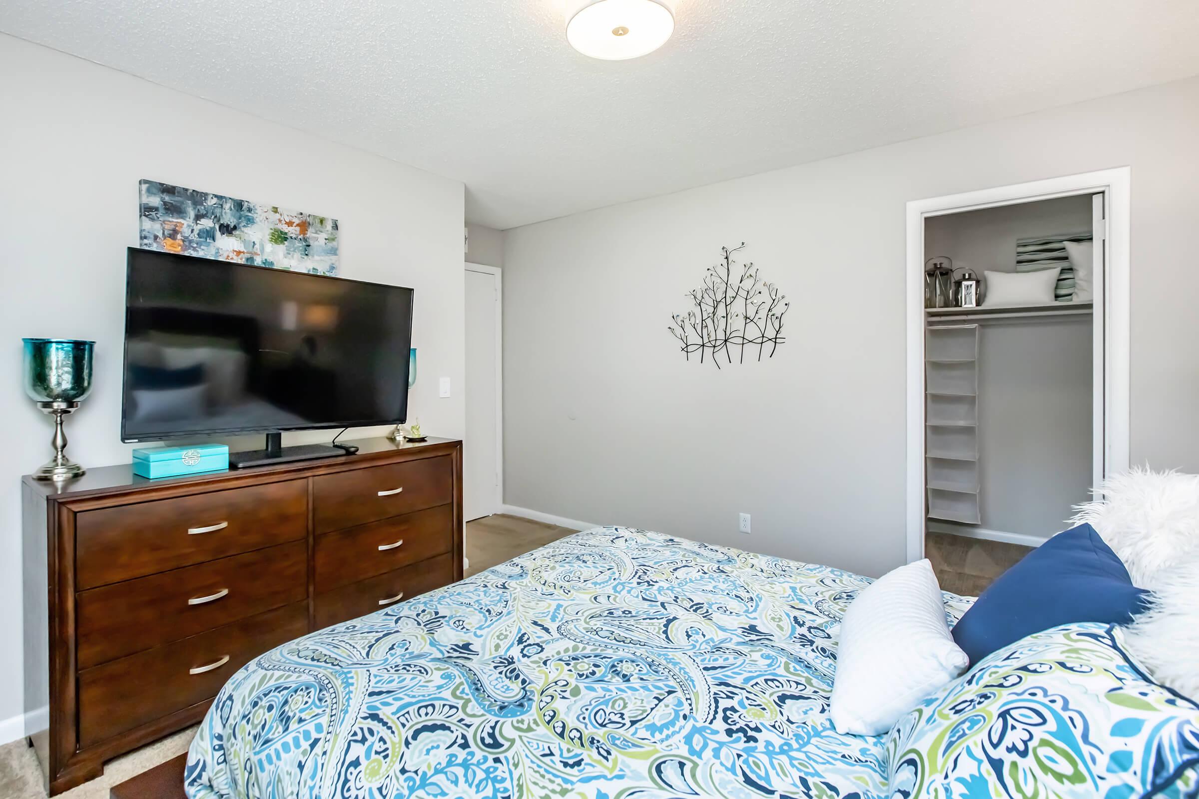 A cozy bedroom featuring a large bed with a decorative blue and green patterned duvet, a dresser with a flat-screen TV on top, and a small accent table. The walls are painted light gray, with a decorative wall art piece and a minimalist closet visible in the background. The room is well-lit and inviting.