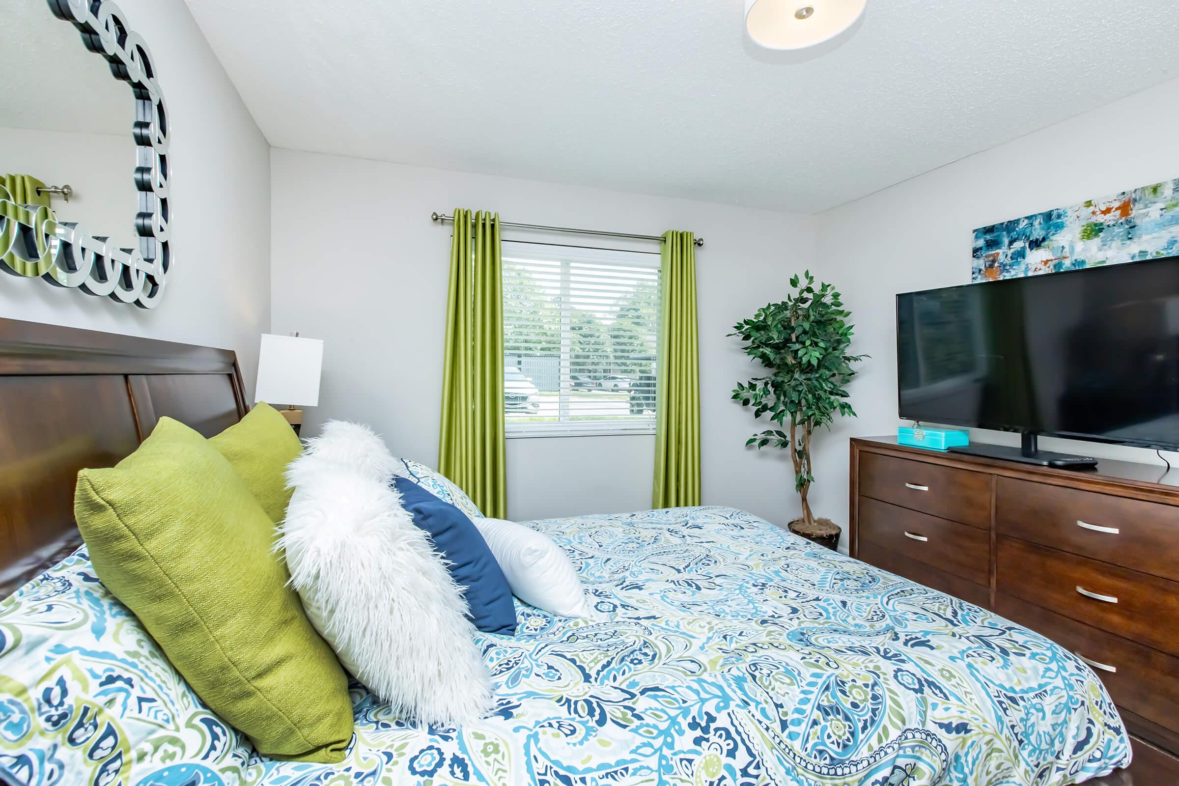 A cozy bedroom featuring a wooden bed with colorful patterned bedding, large pillows, and a fluffy white throw. A green leafy plant stands beside the bed, and a dresser is visible with a flat-screen TV on top. Light streams through a window adorned with green curtains, enhancing the inviting atmosphere.