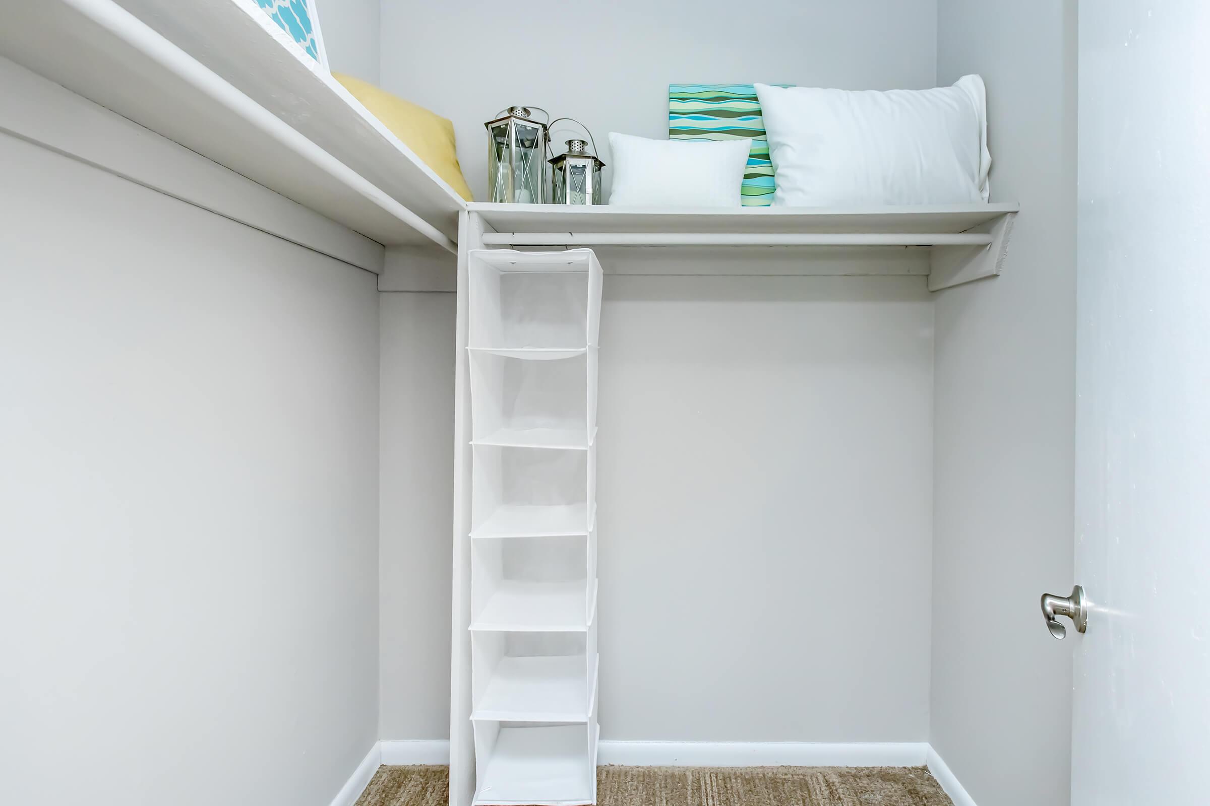 A tidy closet space featuring a high shelf with pillows and a light-colored throw, accompanied by decorative lanterns. A white fabric shelf organizer is positioned on the floor, offering additional storage. The walls are painted a soft gray, and a door is visible on the right side.
