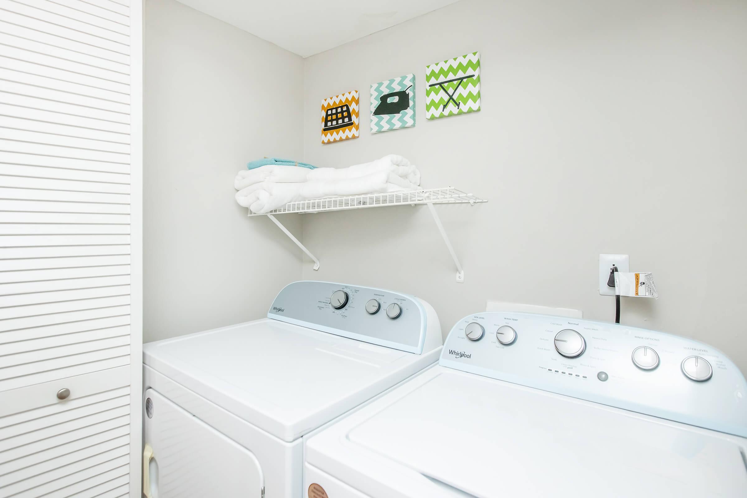 A laundry room featuring a stacked washer and dryer, neatly arranged with white towels on a shelf above. Above the shelf, three colorful framed art pieces add a decorative touch to the gray wall. A white cabinet door is partially visible on the left.