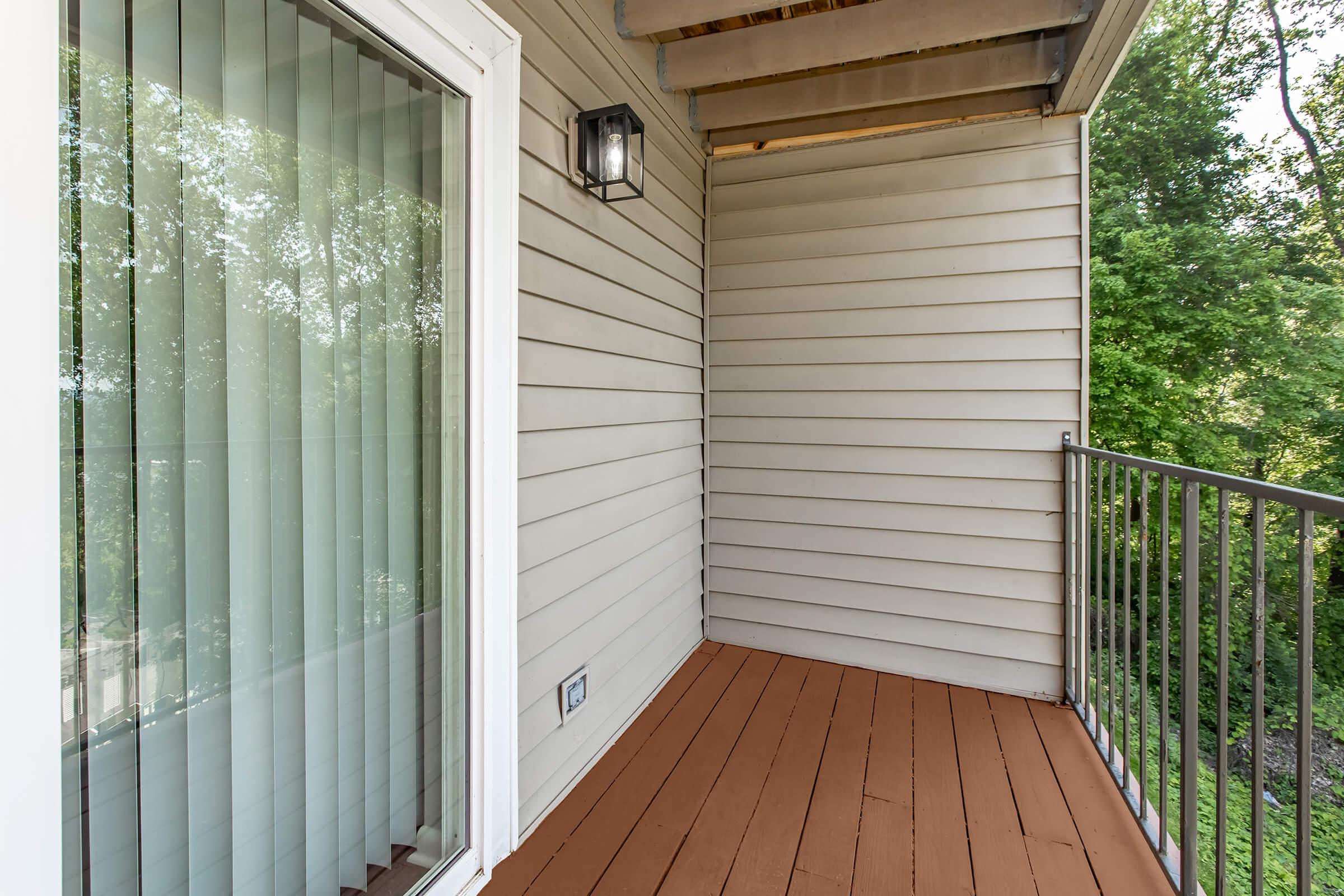 A small balcony with wooden flooring, a gray wall, and large vertical blinds covering a window. There is a modern light fixture on the wall, and green trees are visible outside the railing. The space is well-lit and offers a tranquil outdoor view.