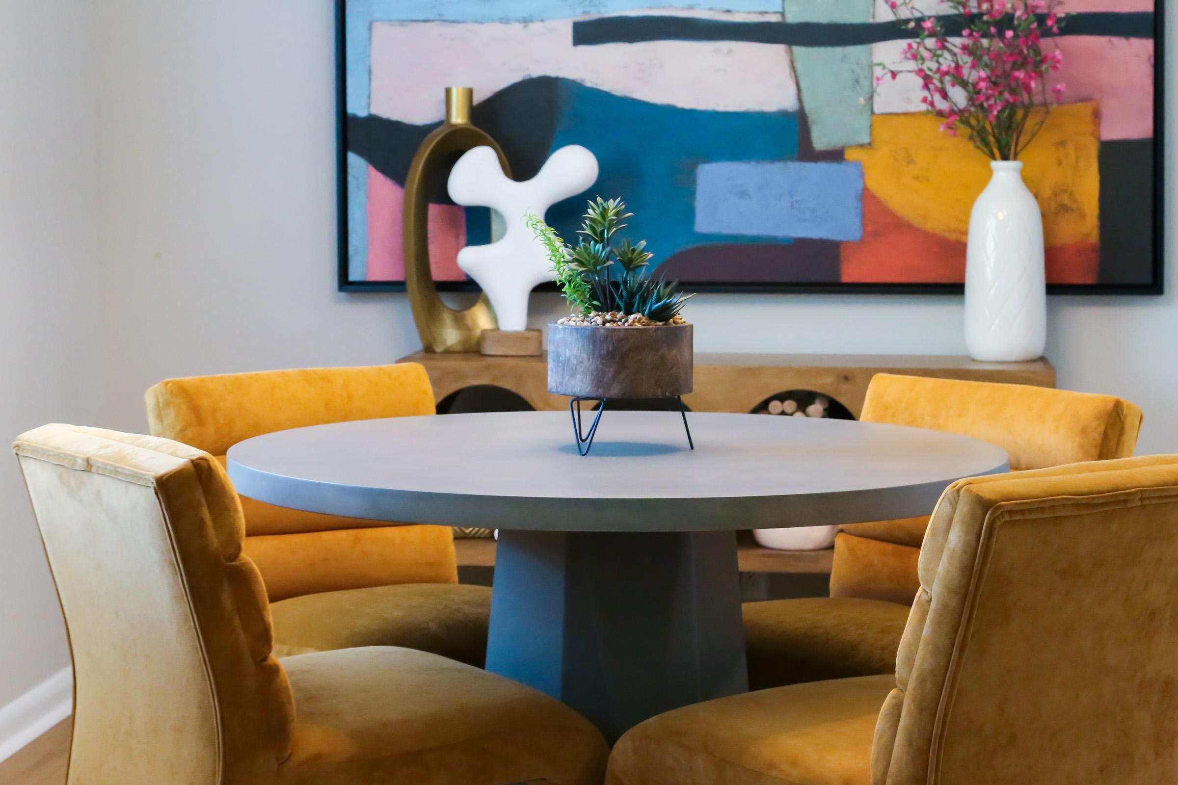 A modern dining area featuring a round gray table with a small, stylish planter in the center. Surrounding the table are four yellow chairs with a plush texture. In the background, colorful abstract artwork adds a vibrant touch, along with a decorative vase and a sculptural piece on a wooden sideboard.