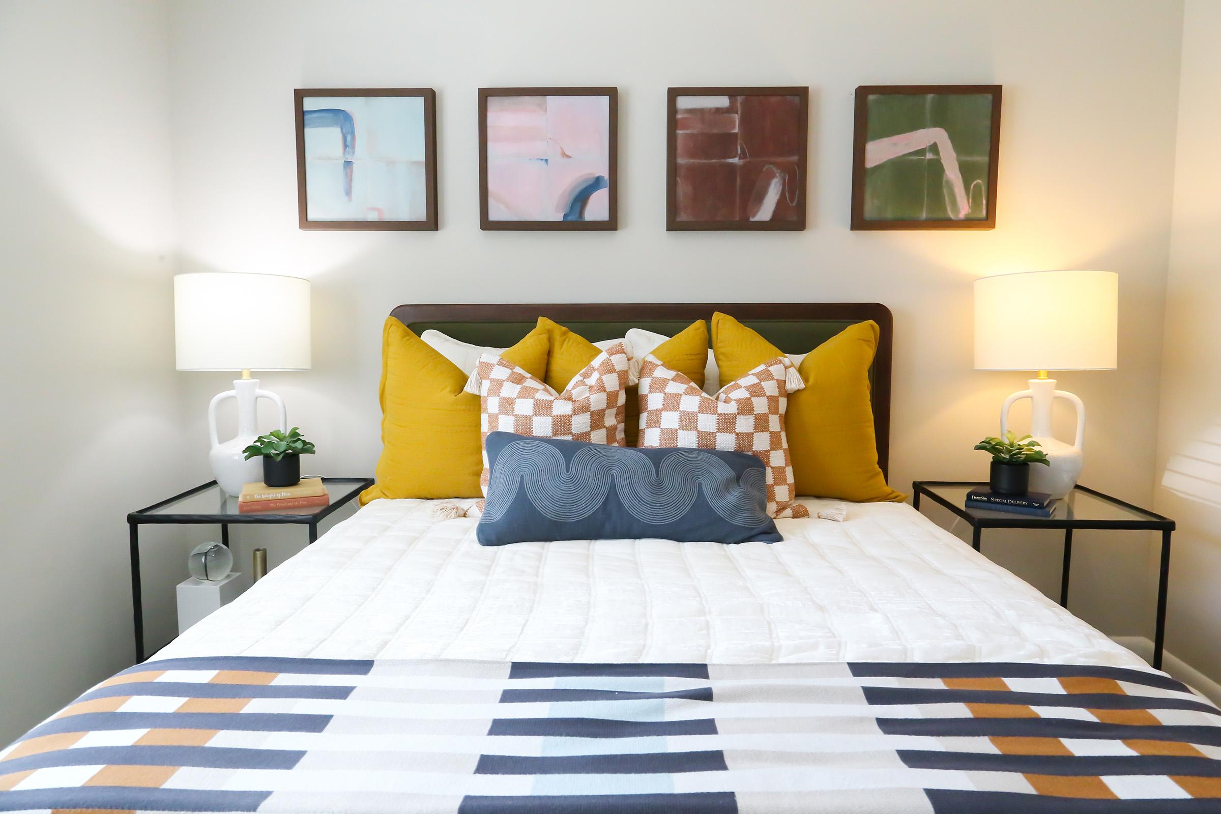A cozy bedroom featuring a neatly made bed with an assortment of decorative pillows in various colors and patterns. Beside the bed, two modern nightstands hold small lamps and potted plants. Above the bed, four abstract art pieces are hung on the wall, adding a stylish touch to the room.