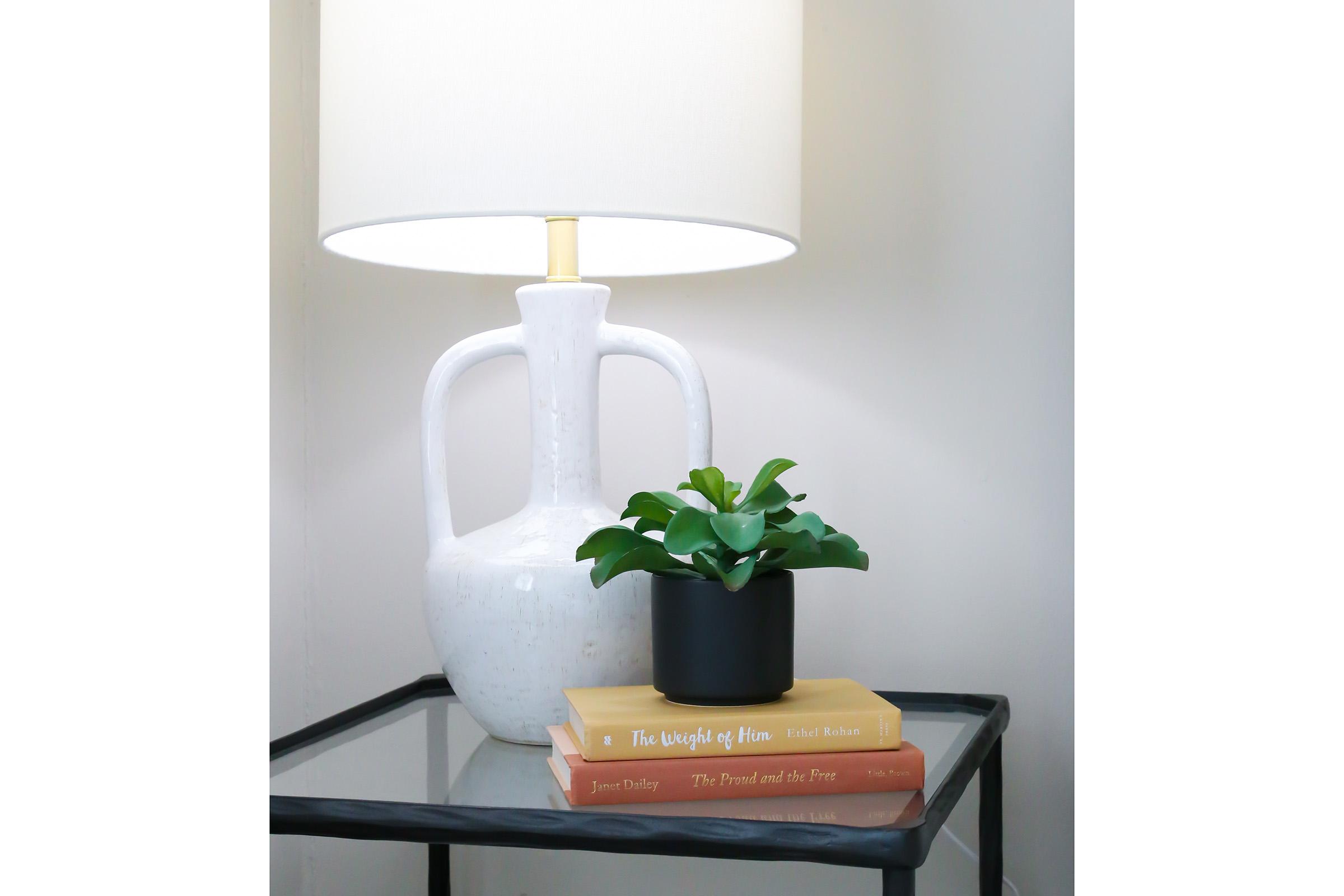 A stylish table featuring a white ceramic lamp with a round shade, a stack of books in warm colors, and a small green plant in a black pot. The setting is minimalistic and modern, with a glass top table enhancing the aesthetic.