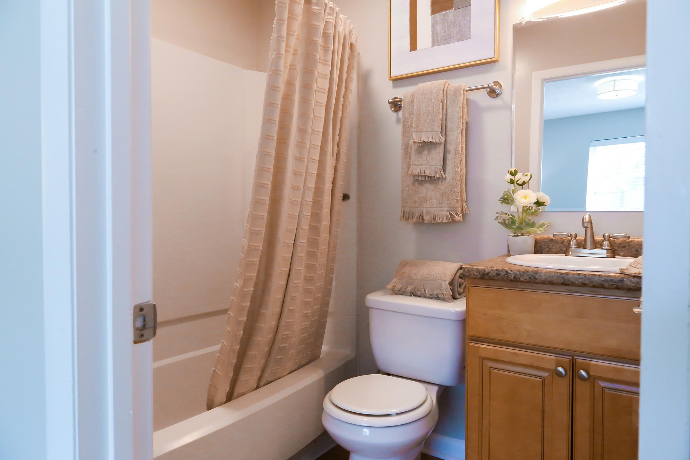 A well-lit bathroom featuring a shower with a beige curtain, a white toilet, and a vanity with a sink. The vanity has a granite countertop adorned with a folded towel and a small vase with flowers. Decorative towels are hanging on a rack, and a framed artwork is on the wall.